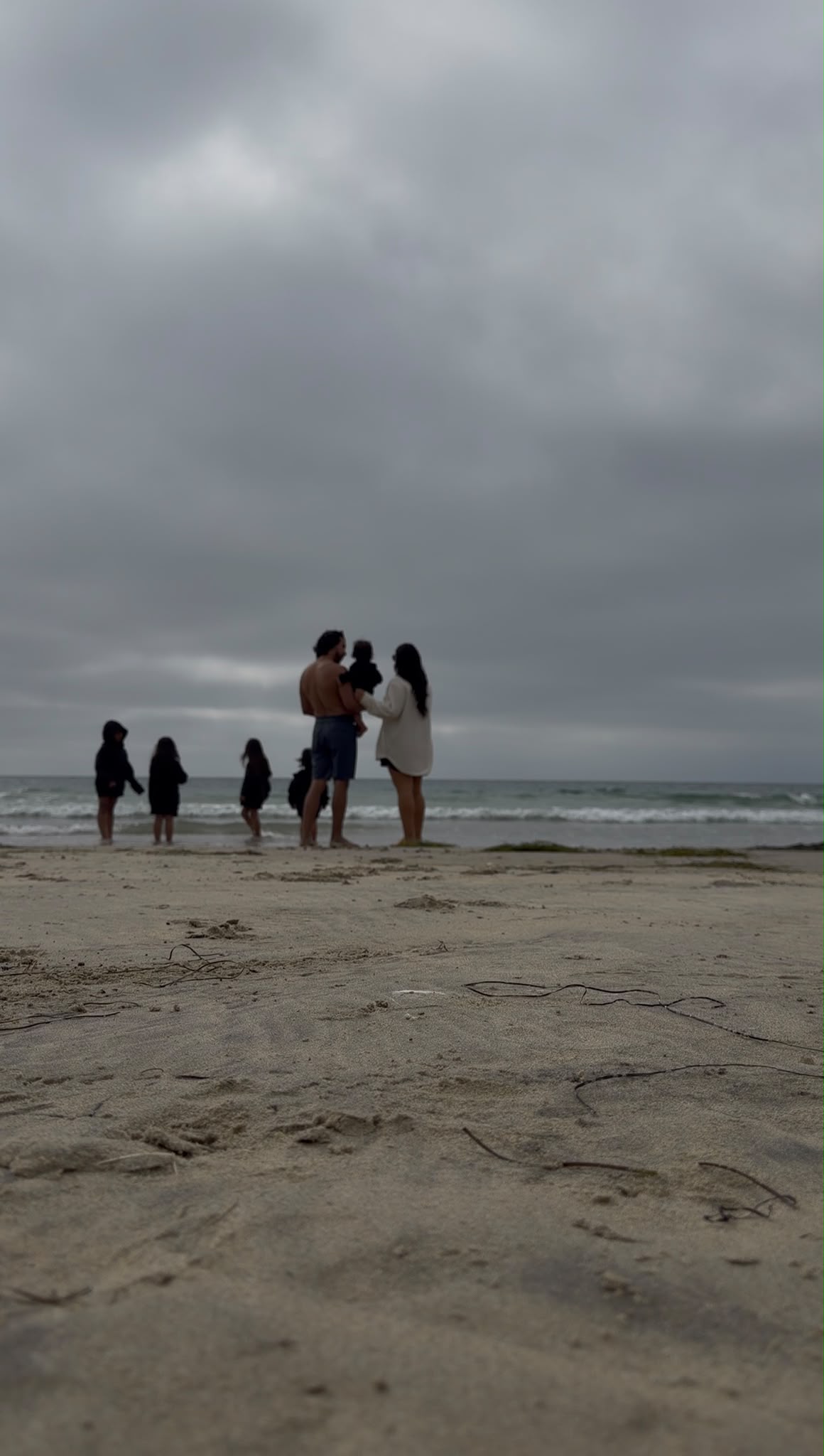 Group of people standing on a beach under cloudy sky, some talking and others walking near the water.