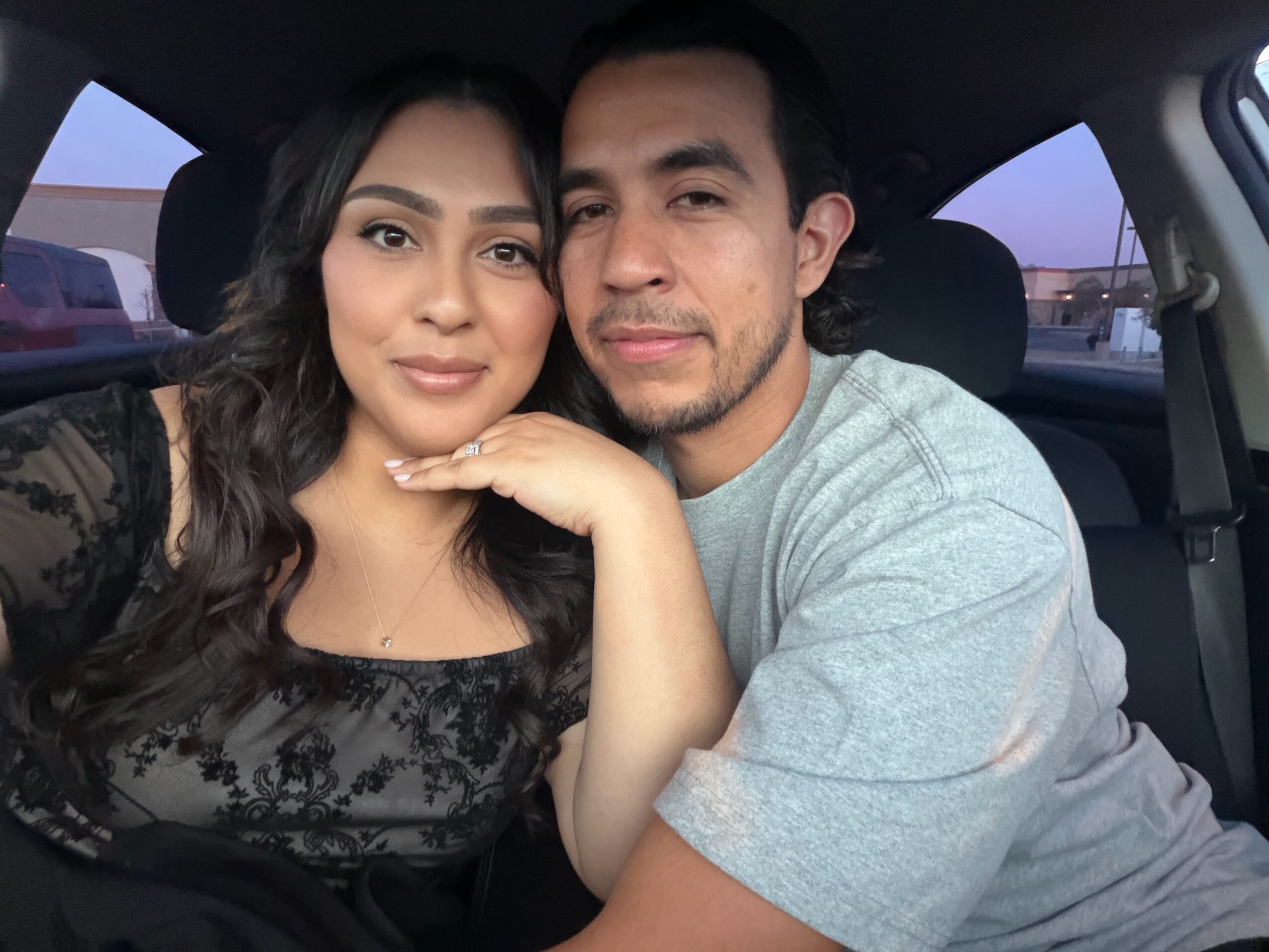 A woman and a man sit close together inside a car, smiling at the camera, with sunset visible through the window.