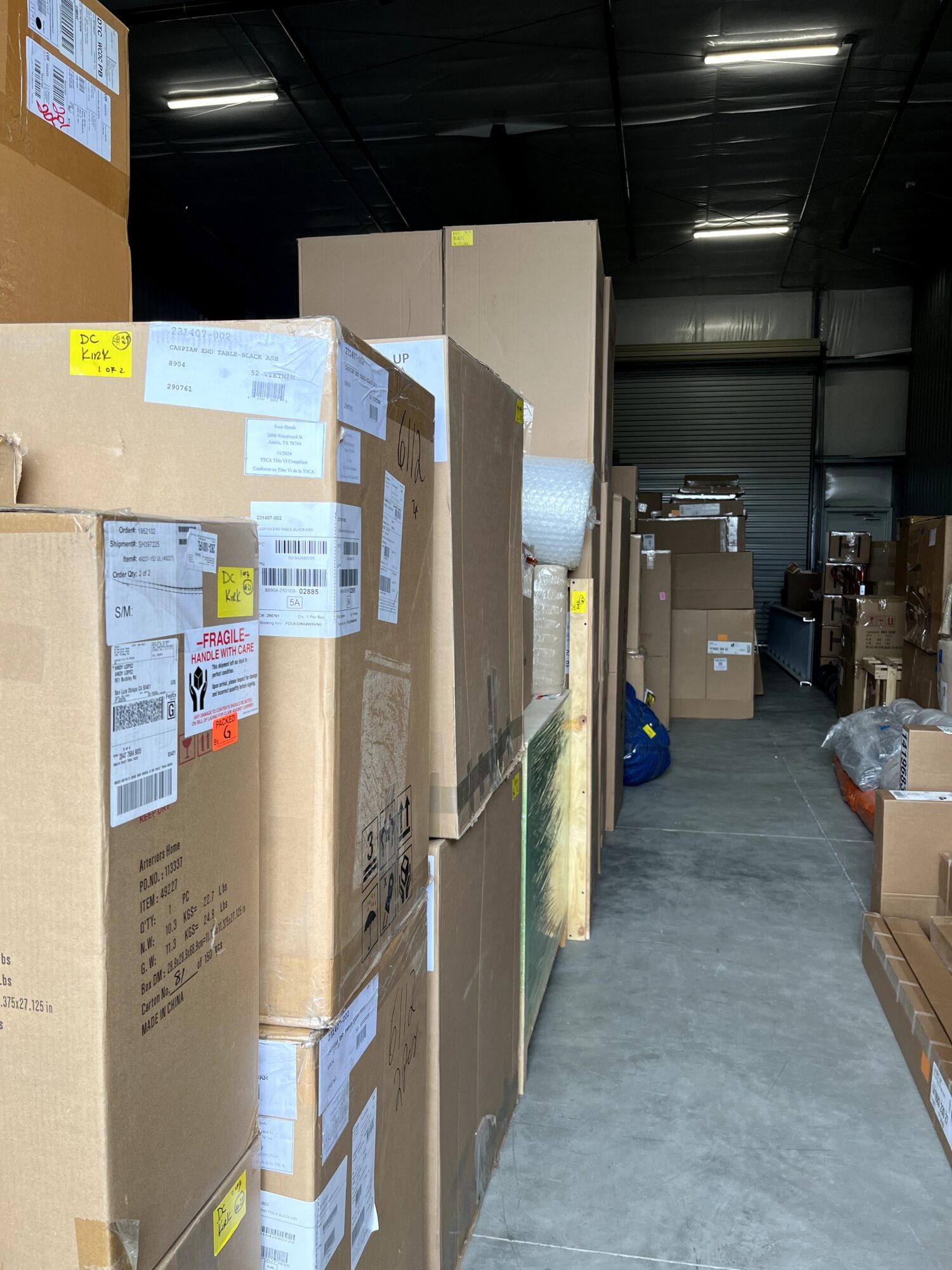 Stacked cardboard boxes in a warehouse with a concrete floor and dark ceiling, some labels visible on boxes.