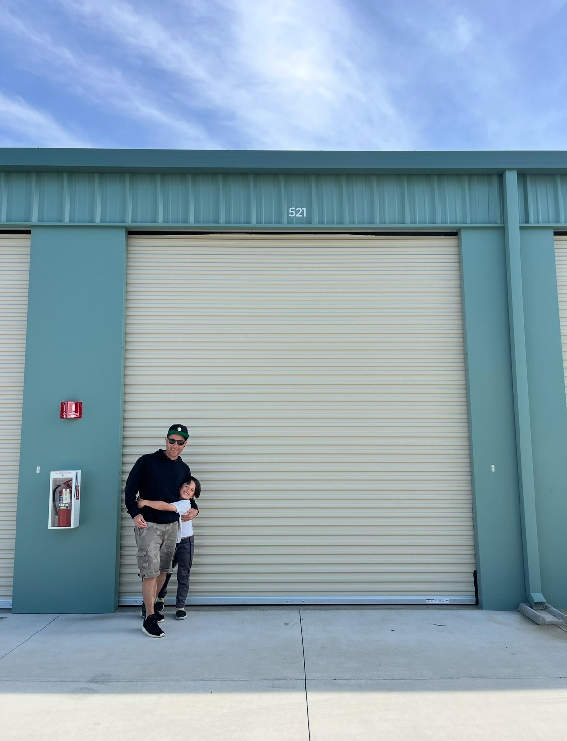 Two people stand in front of a large closed garage door on a teal building, under a partly cloudy sky.