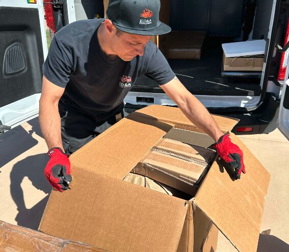 Man wearing a black cap and gloves, packing boxes into a van, with open van door behind him.