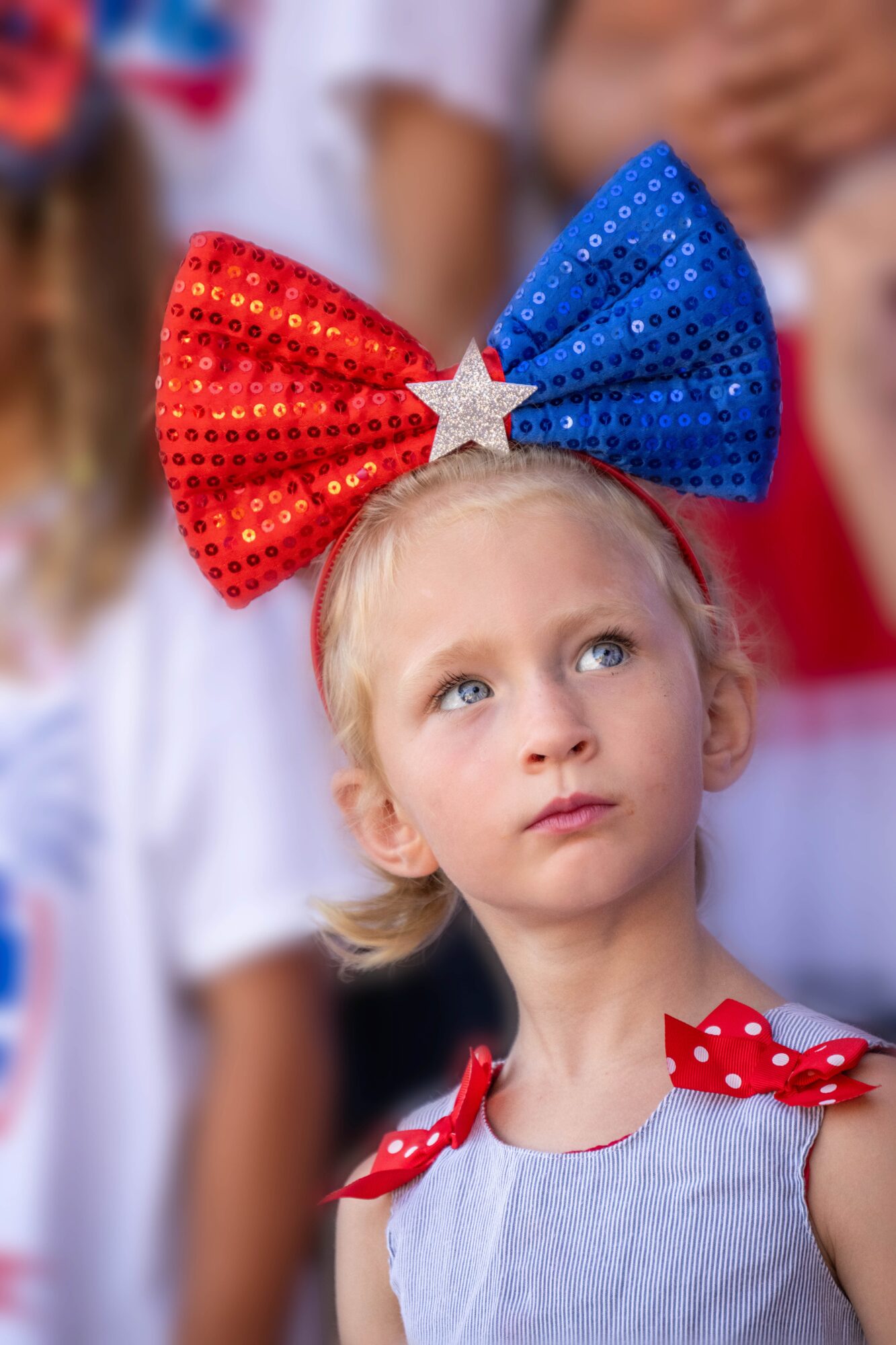 Young girl with a large red and blue sequin bow headband with a star, looking upward, wearing a light-colored dress with red polka-dot bows on shoulders.