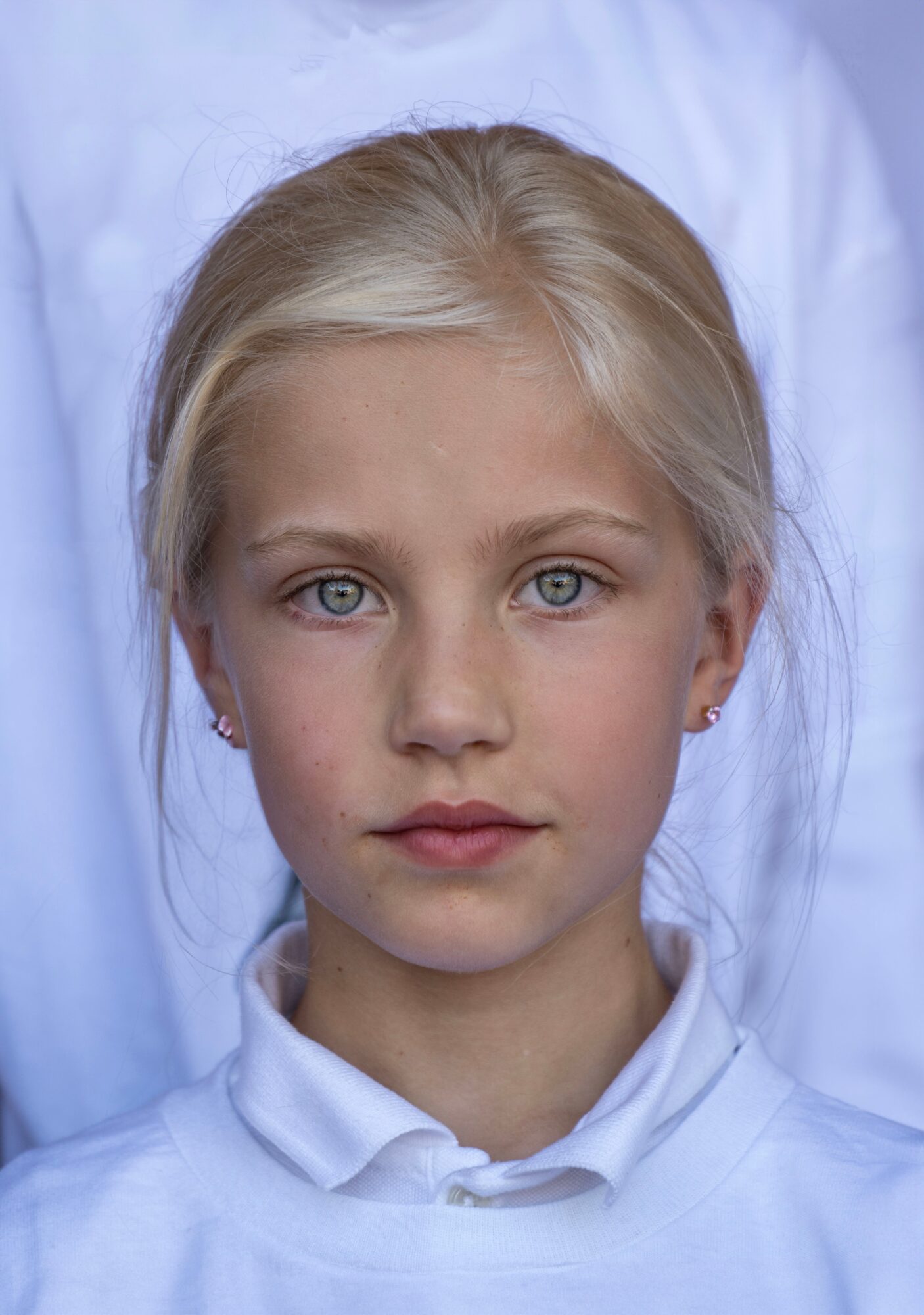 A young girl with blonde hair and blue eyes, wearing a white shirt, looking directly at the camera.