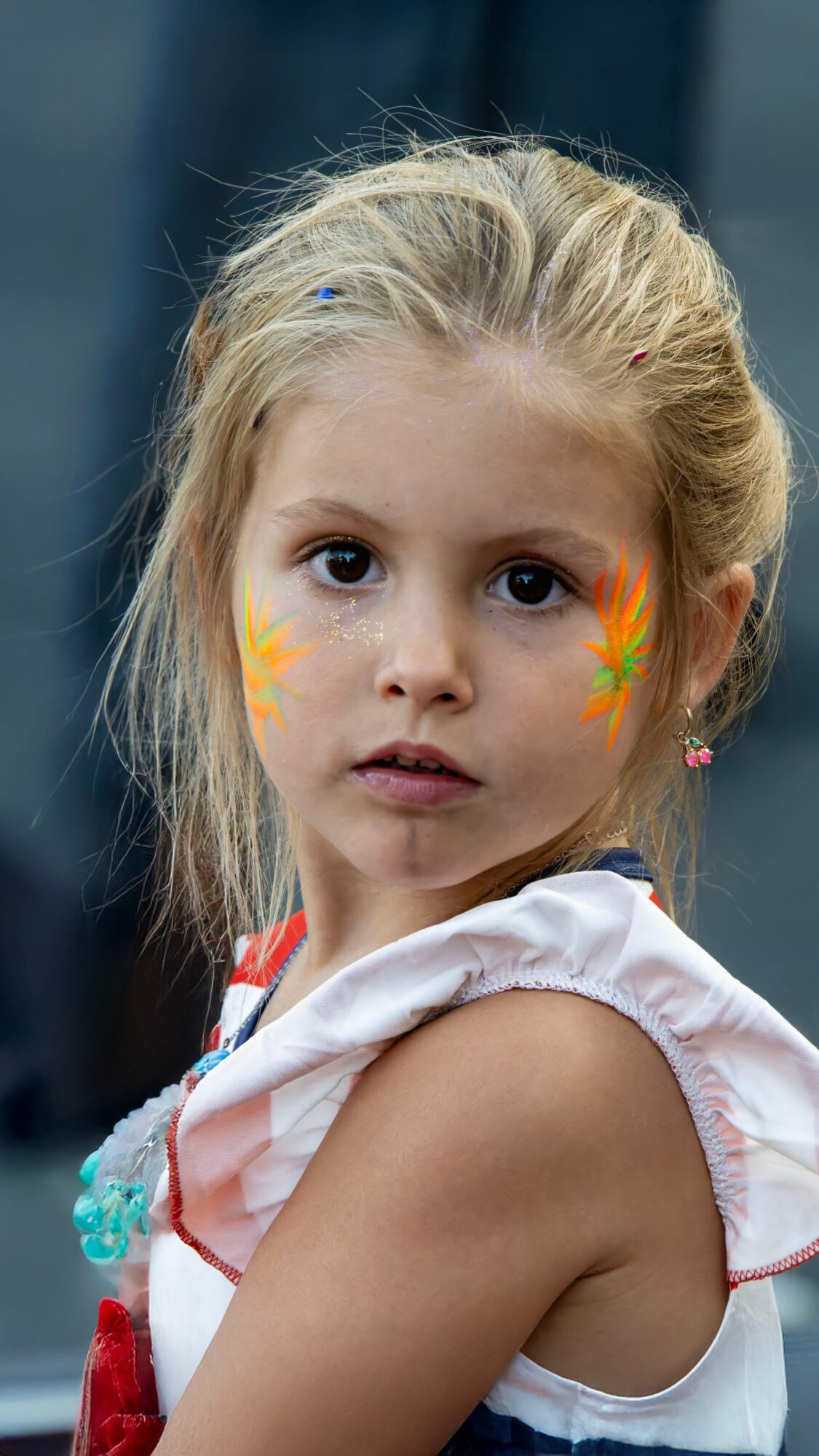 Young girl with face paint, wearing a white top, looking at the camera outdoors.