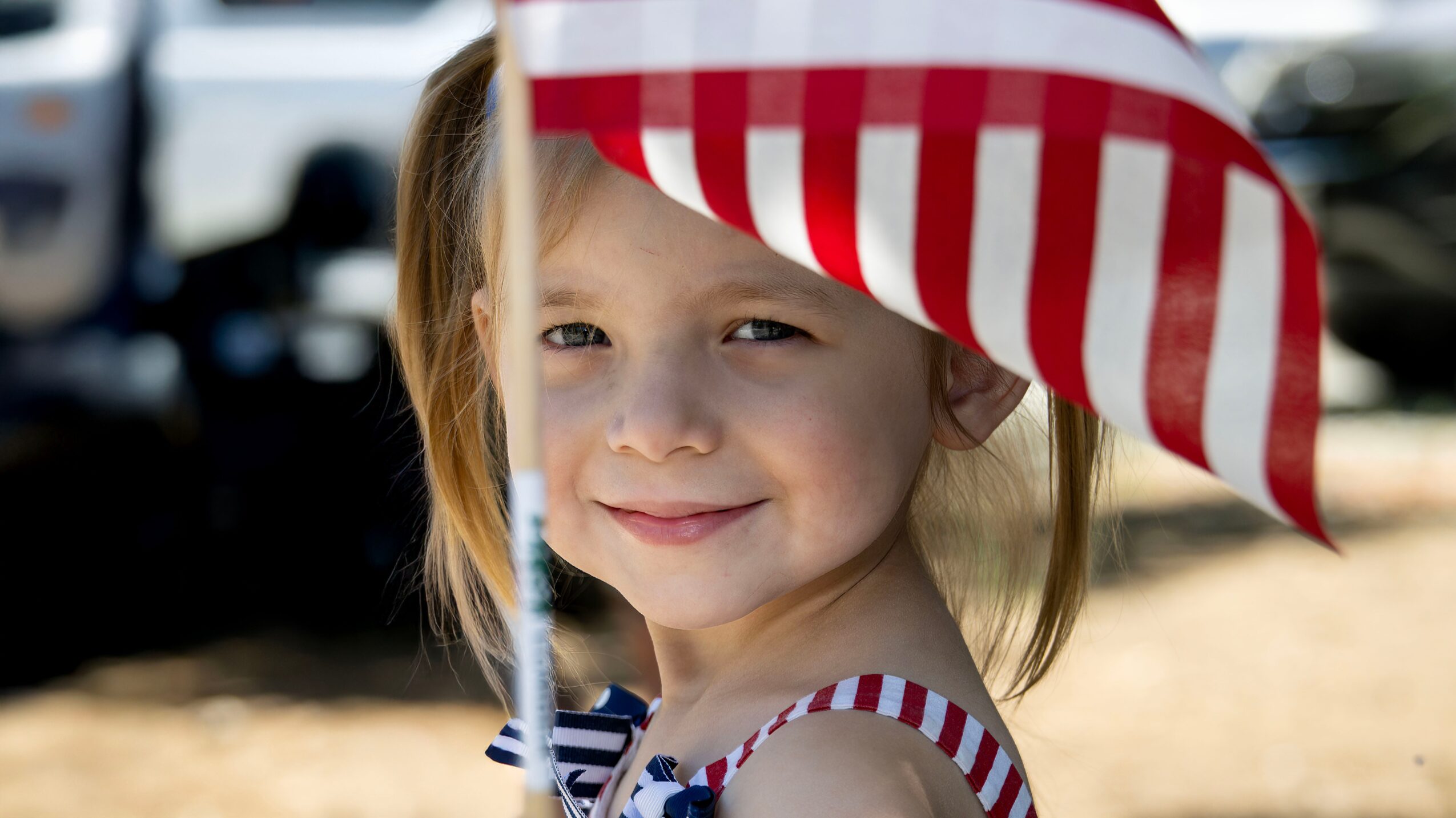Young girl smiling outdoors holding a red and white striped umbrella, sunlight on her face, blurred background.