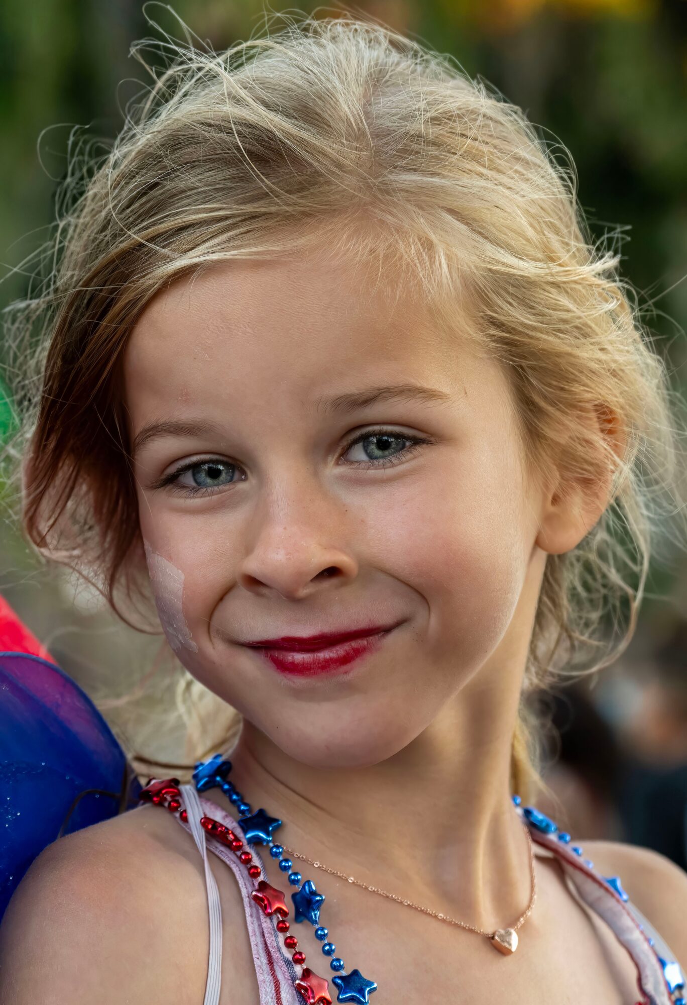 Young girl with blonde hair and blue eyes smiling outdoors, wearing a colorful necklace and a light top.
