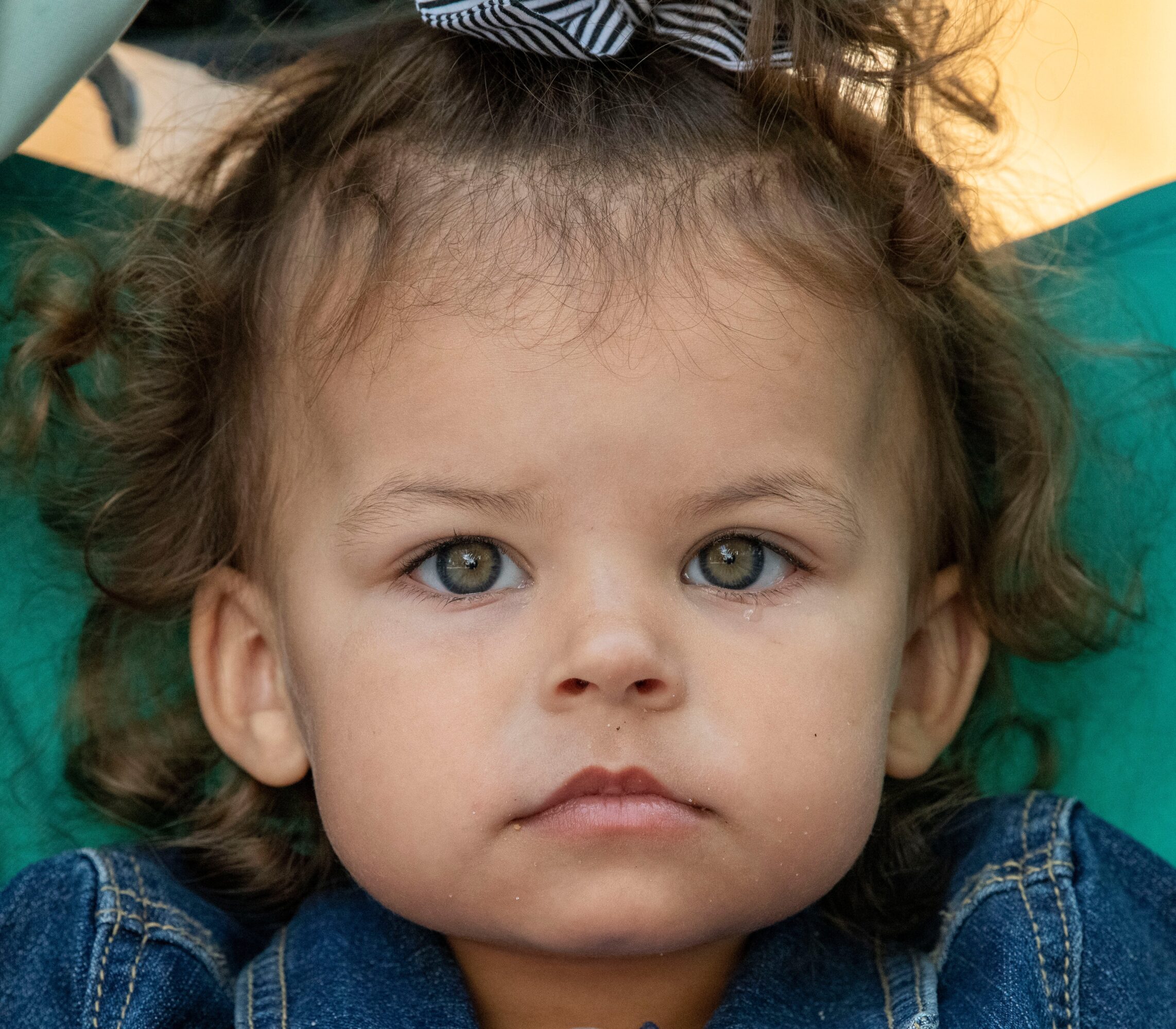 Close-up of a young girl with curly hair, blue eyes, and a striped bow on her head, wearing a denim jacket.