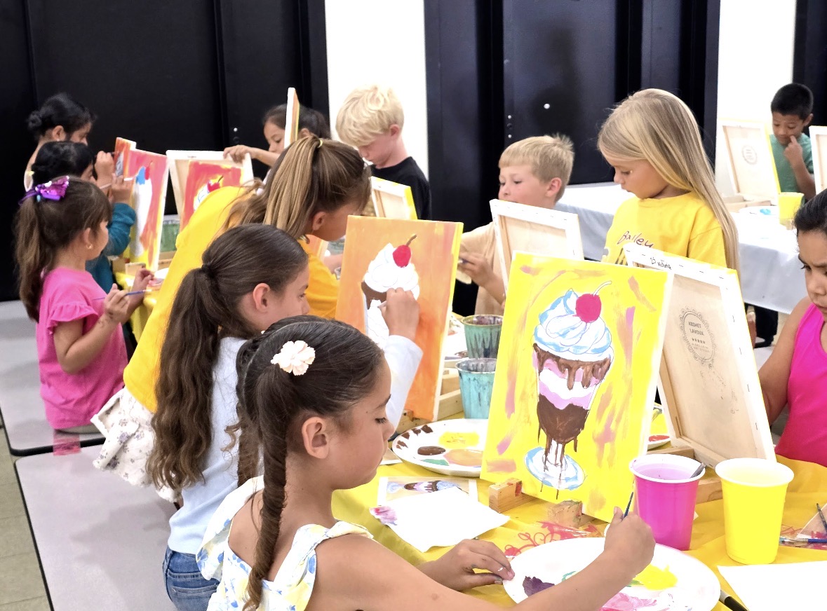 Children painting colorful pictures of desserts at a table in a classroom or art studio.