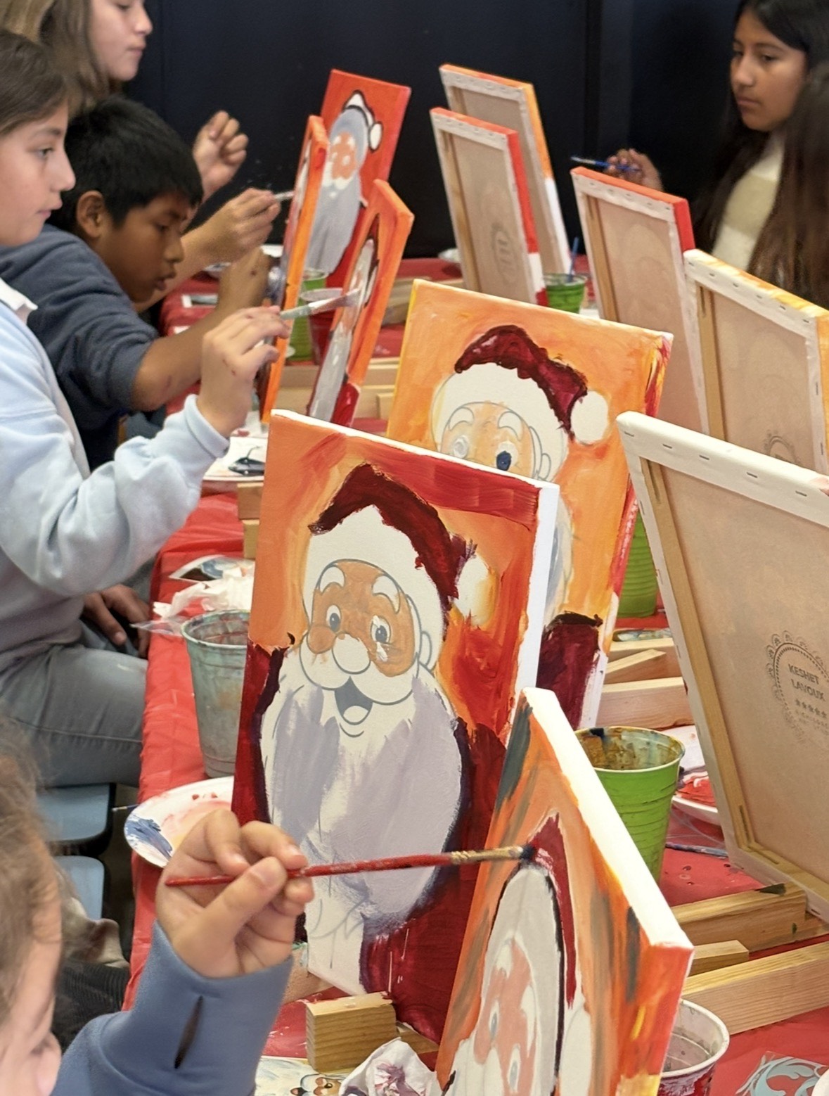 Children painting holiday-themed portraits on canvases at a table, with easels and art supplies visible.