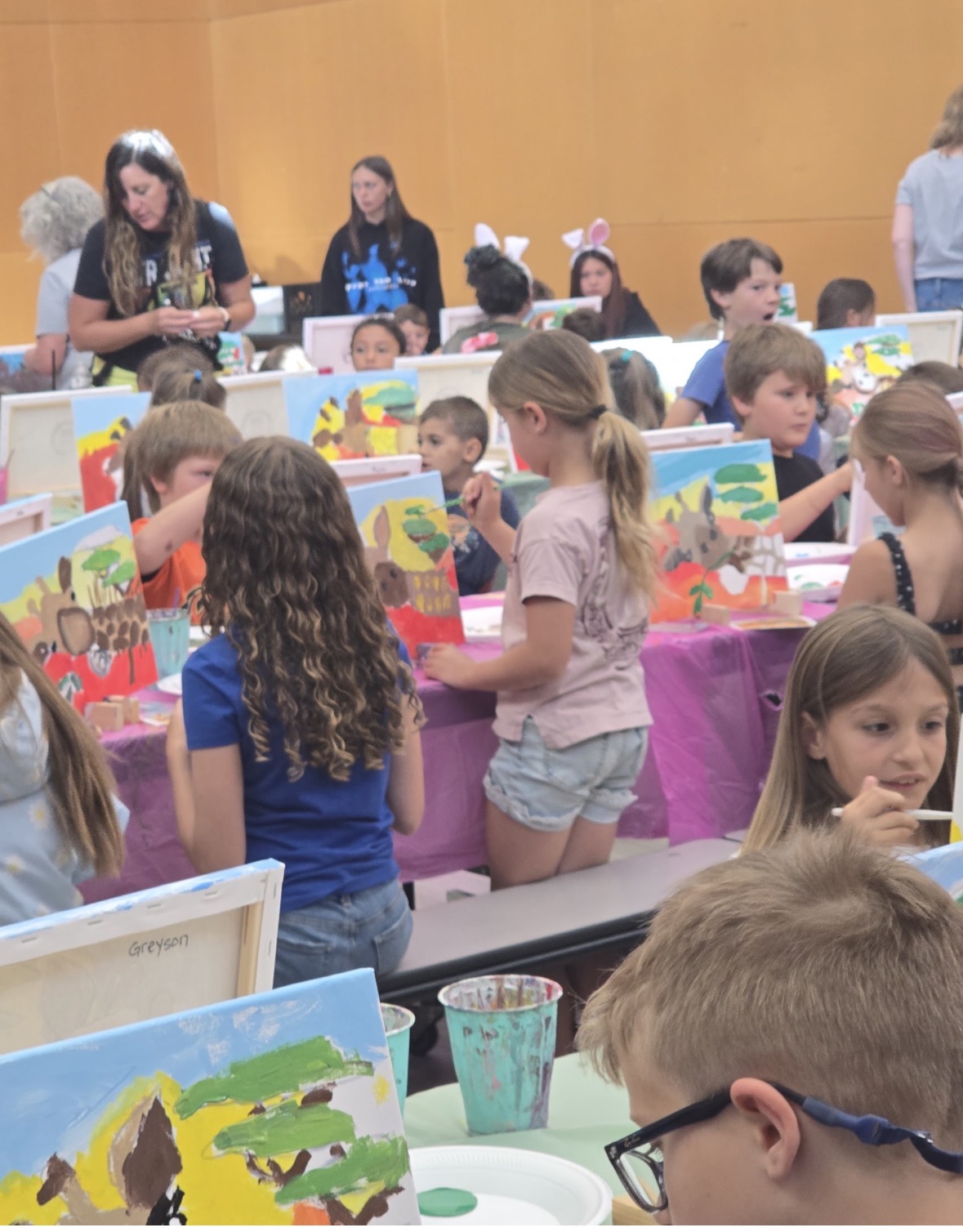 Children gather around tables with colorful artwork, engaging in an art activity in a room with orange walls.
