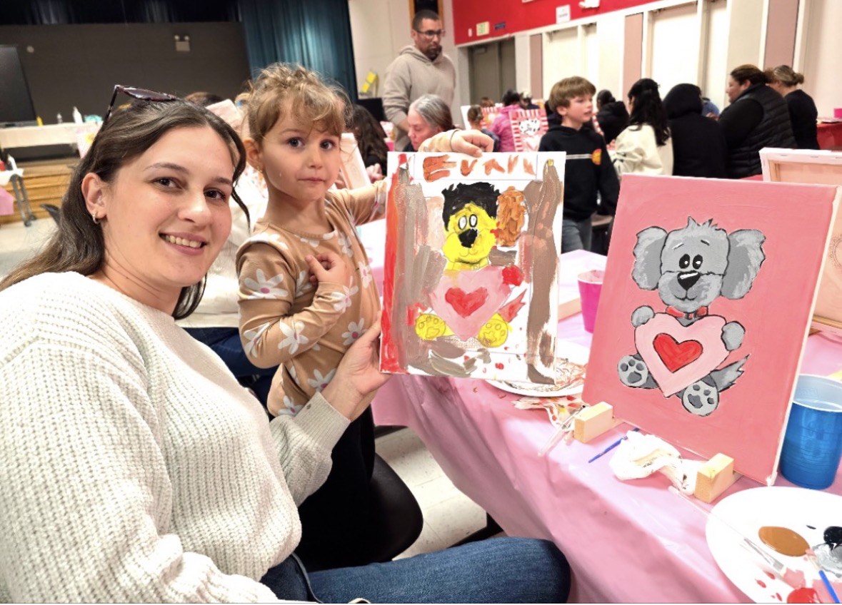 Woman and girl at arts and crafts table with painted canvas artworks, in a busy indoor setting.