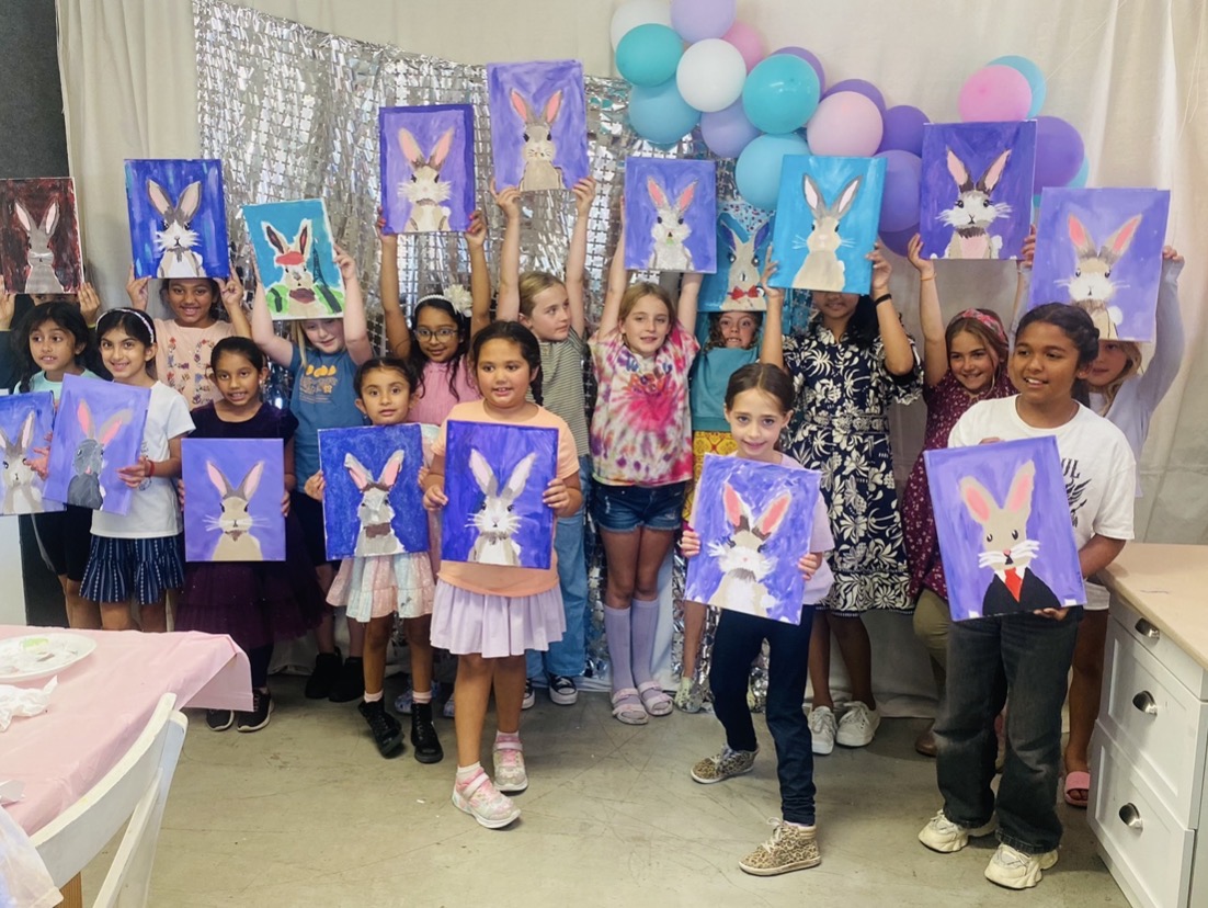 Group of children holding paintings of a bunny with a purple background, decorated with balloons and a shiny backdrop.