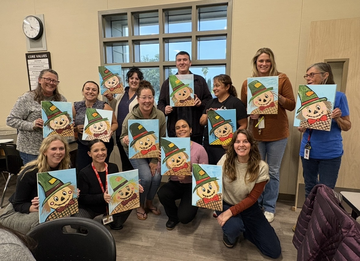 Group of people holding colorful snowman paintings indoors, smiling, with a window and beige wall in background.