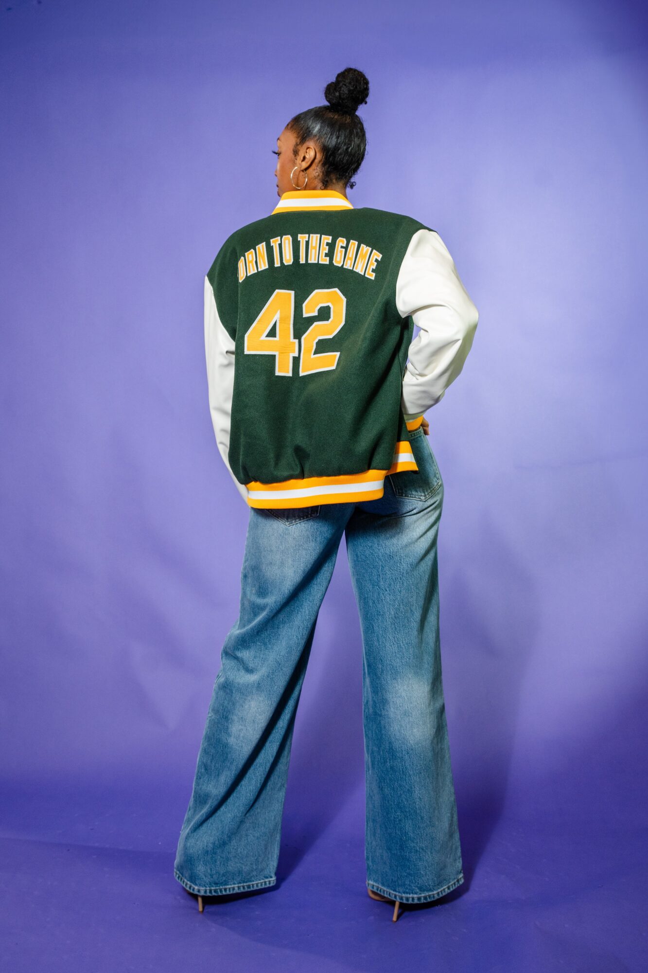 Person with a high bun wearing a green and white varsity jacket with the number 42, facing away, standing against a purple background.