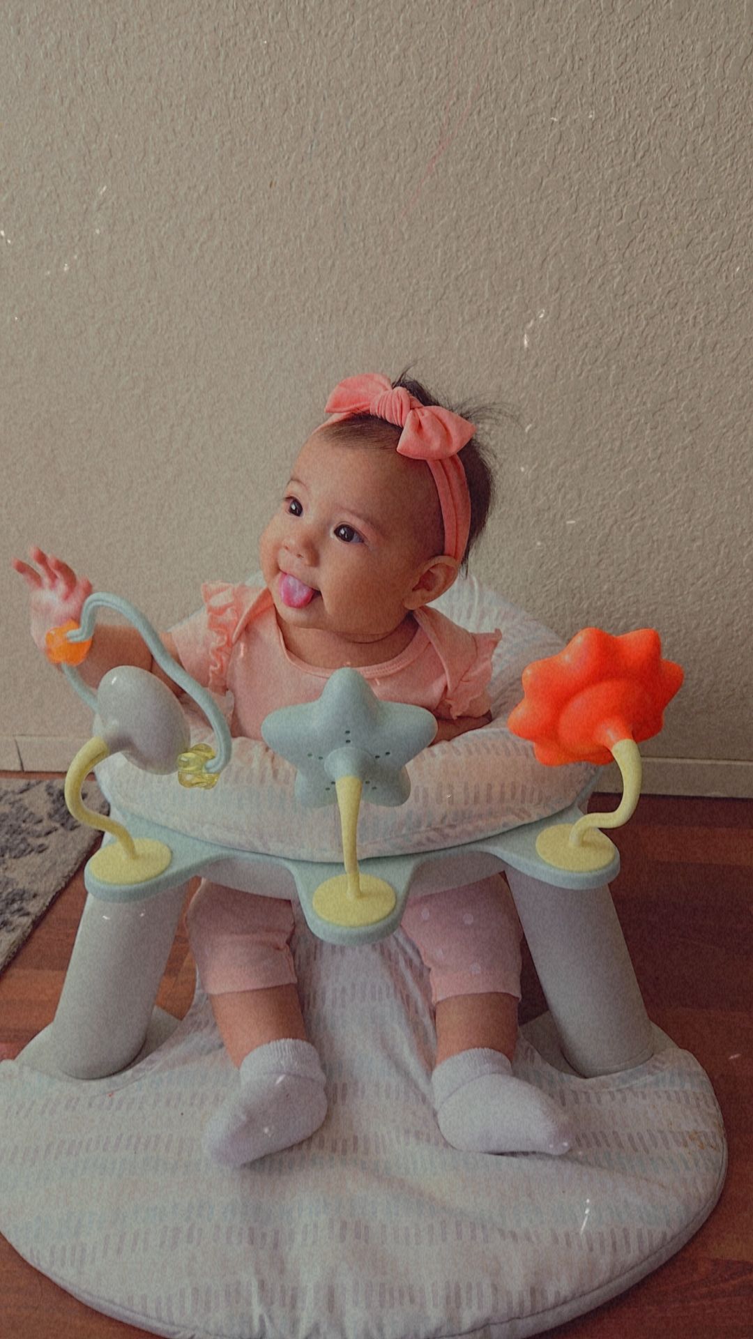 Baby girl with pink bow in hair sitting in a baby jumper with colorful toys, on a carpeted floor.