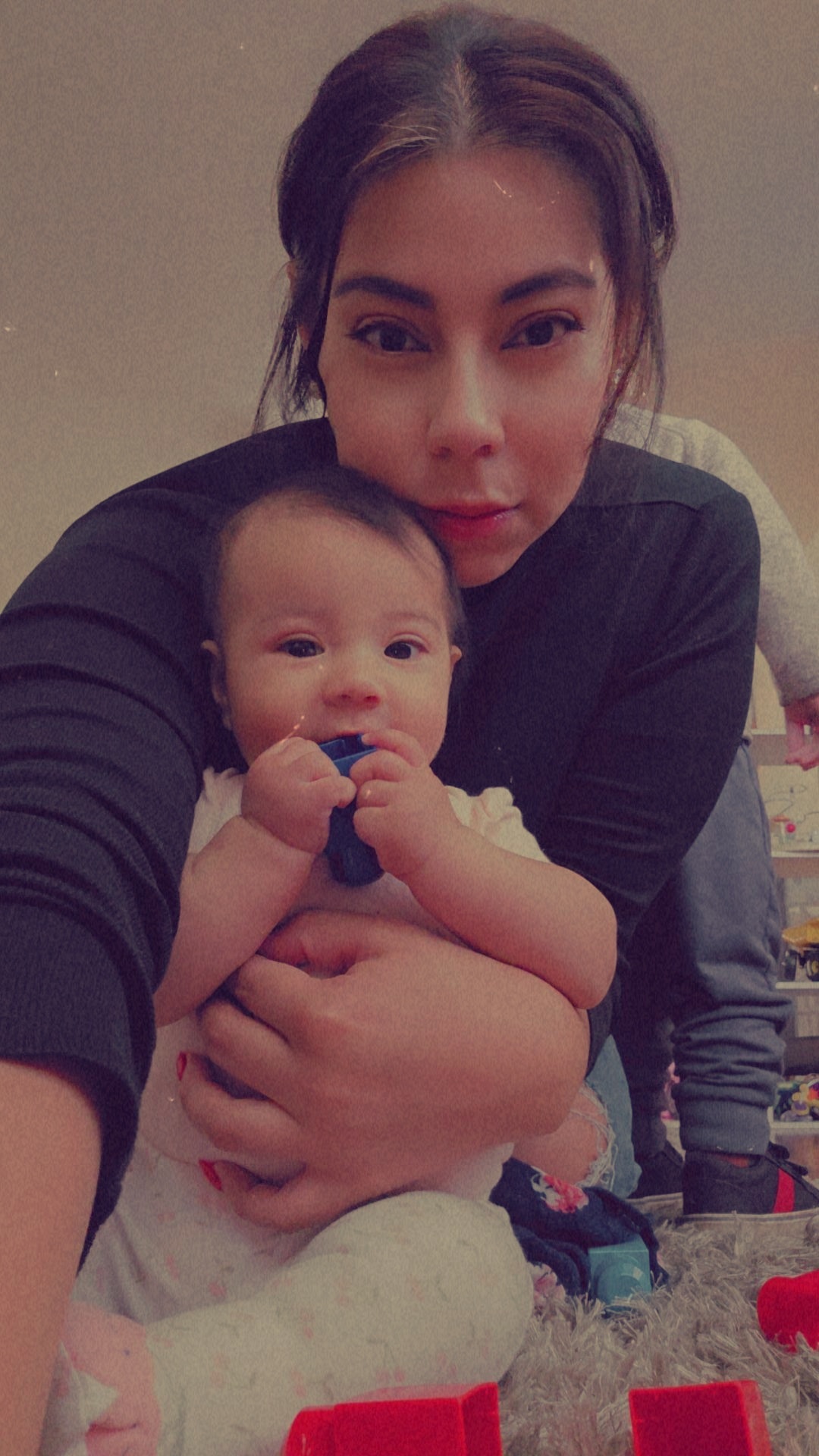 Young woman holding a baby, both looking at the camera, indoors with toys in the background.