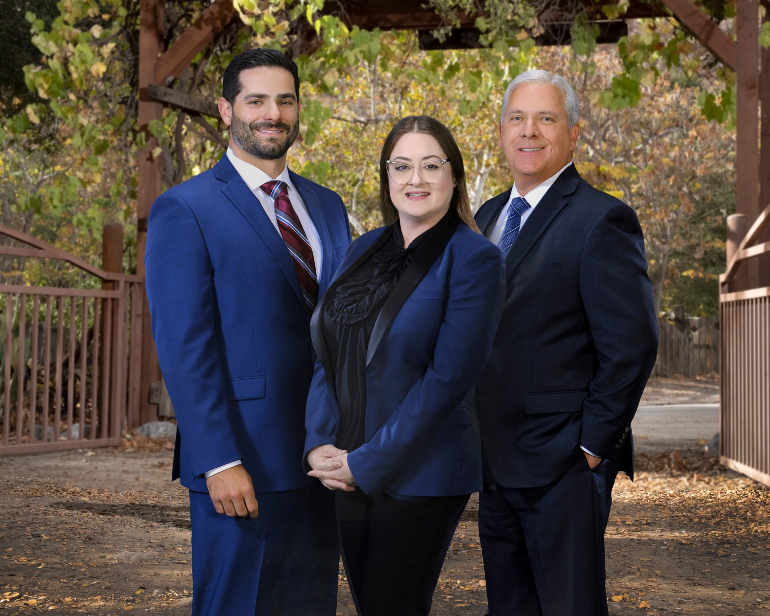 Three people in formal attire standing outdoors under a wooden structure with trees in the background.