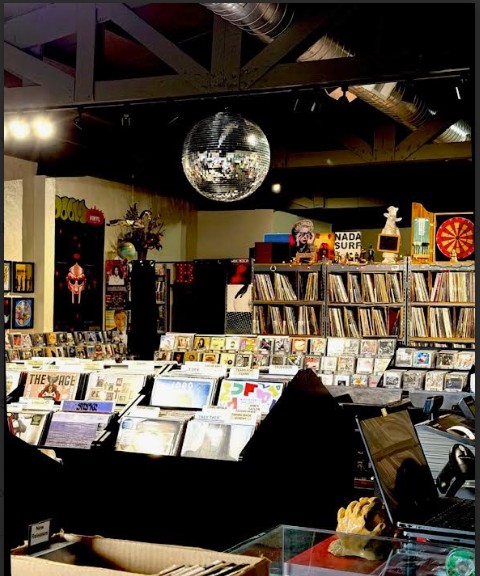Record store with shelves of vinyl records, a disco ball hanging from the ceiling, and a person browsing records.