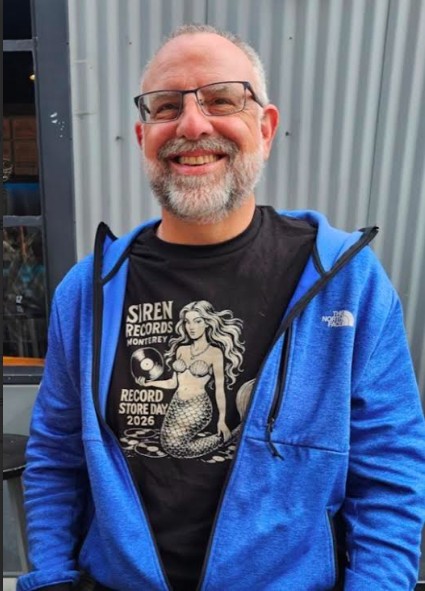 Smiling man with glasses and a beard, wearing a black T-shirt and blue jacket, standing outdoors in front of a metal wall.