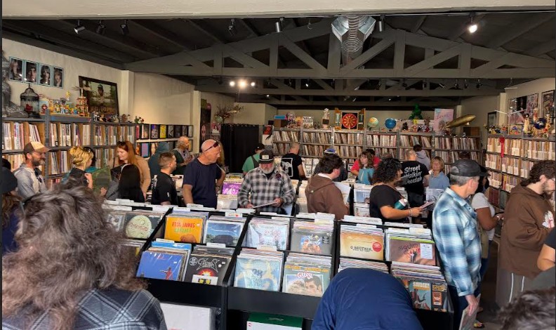People browsing bookshelves and tables in a bookstore or library interior, with shelves filled with books and a high ceiling.