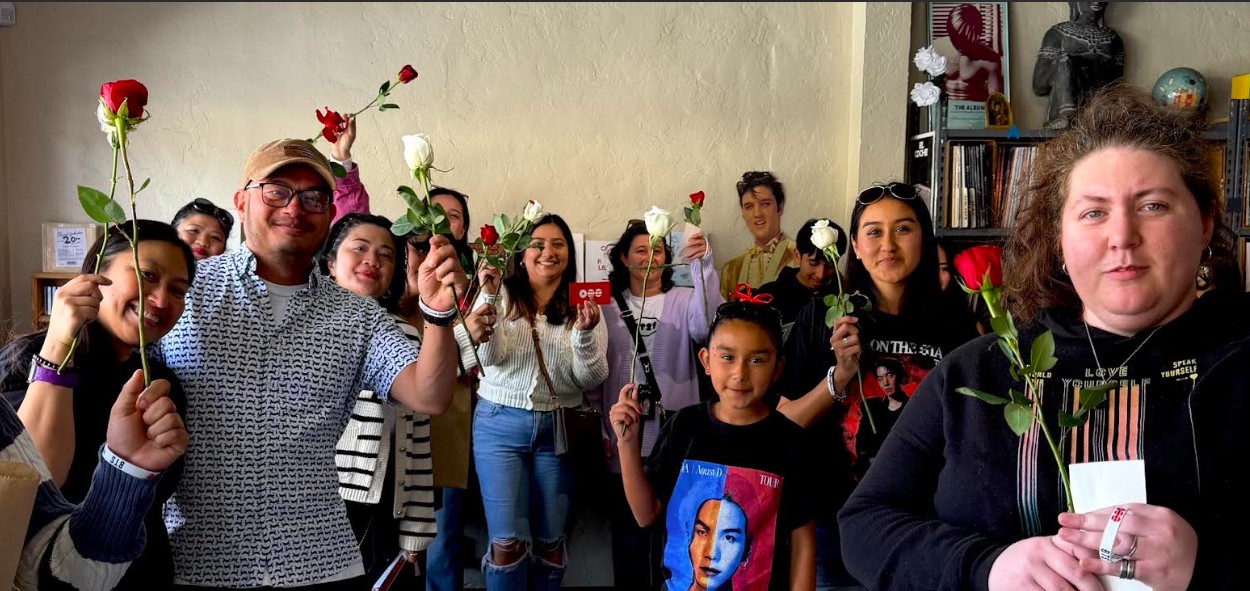 Group of people holding roses indoors, some smiling, some serious, with various ages and backgrounds, in a casual setting.