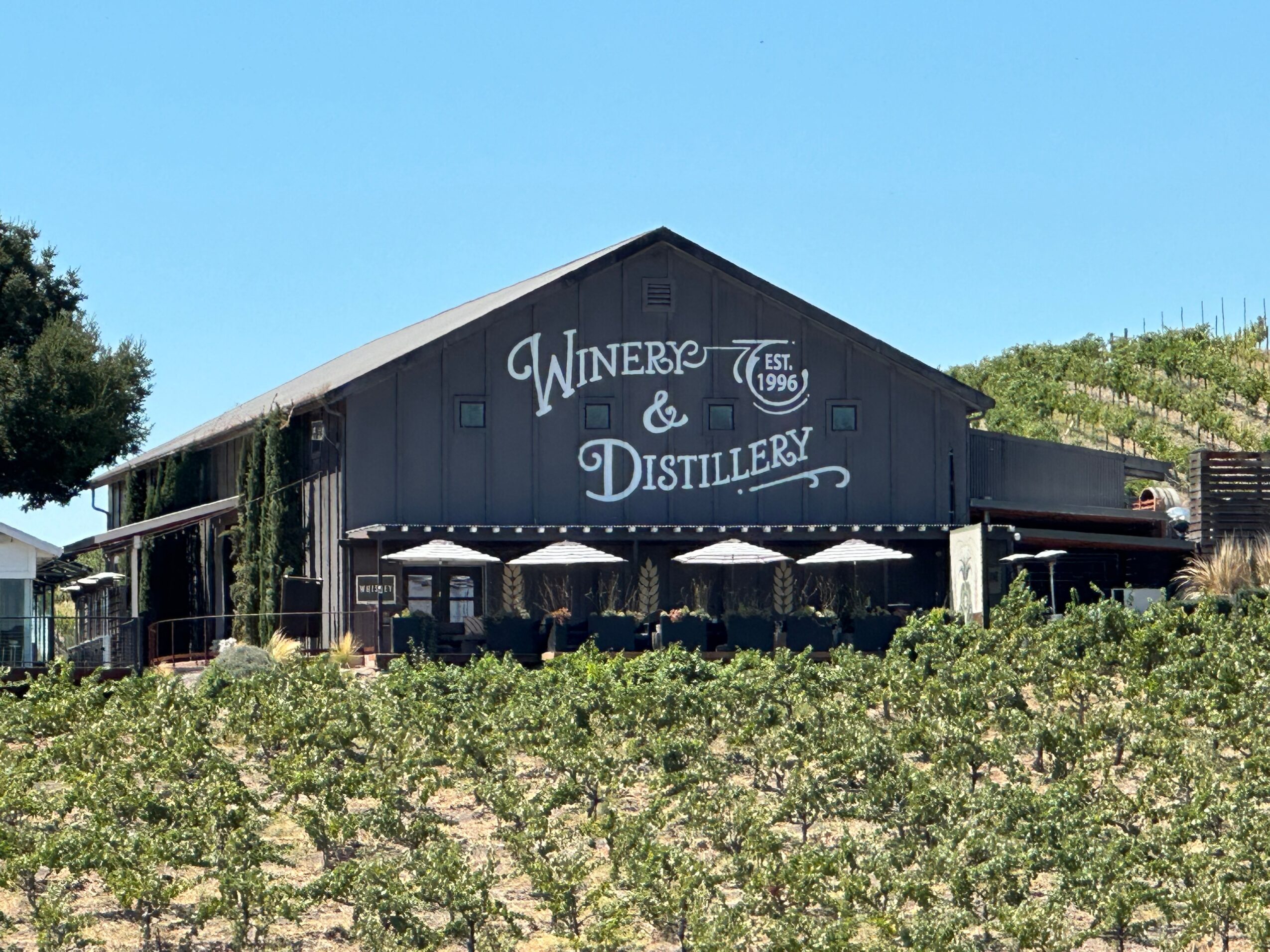 Black building with white sign reading 'Winery & Distillery' under a clear blue sky, surrounded by greenery and outdoor seating with umbrellas.