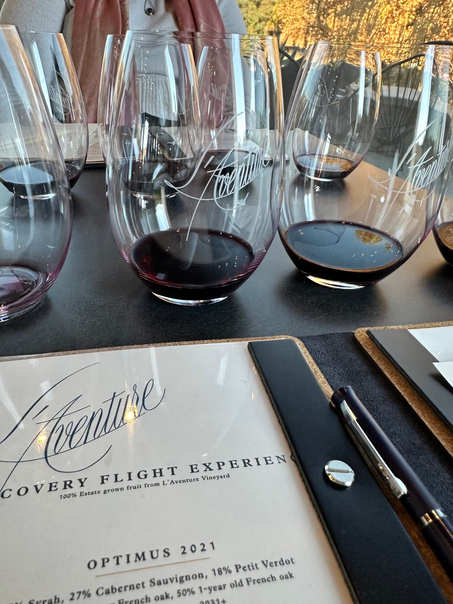 Multiple wine glasses filled with red wine on a table, with a menu and pen nearby, outdoors with trees in background.