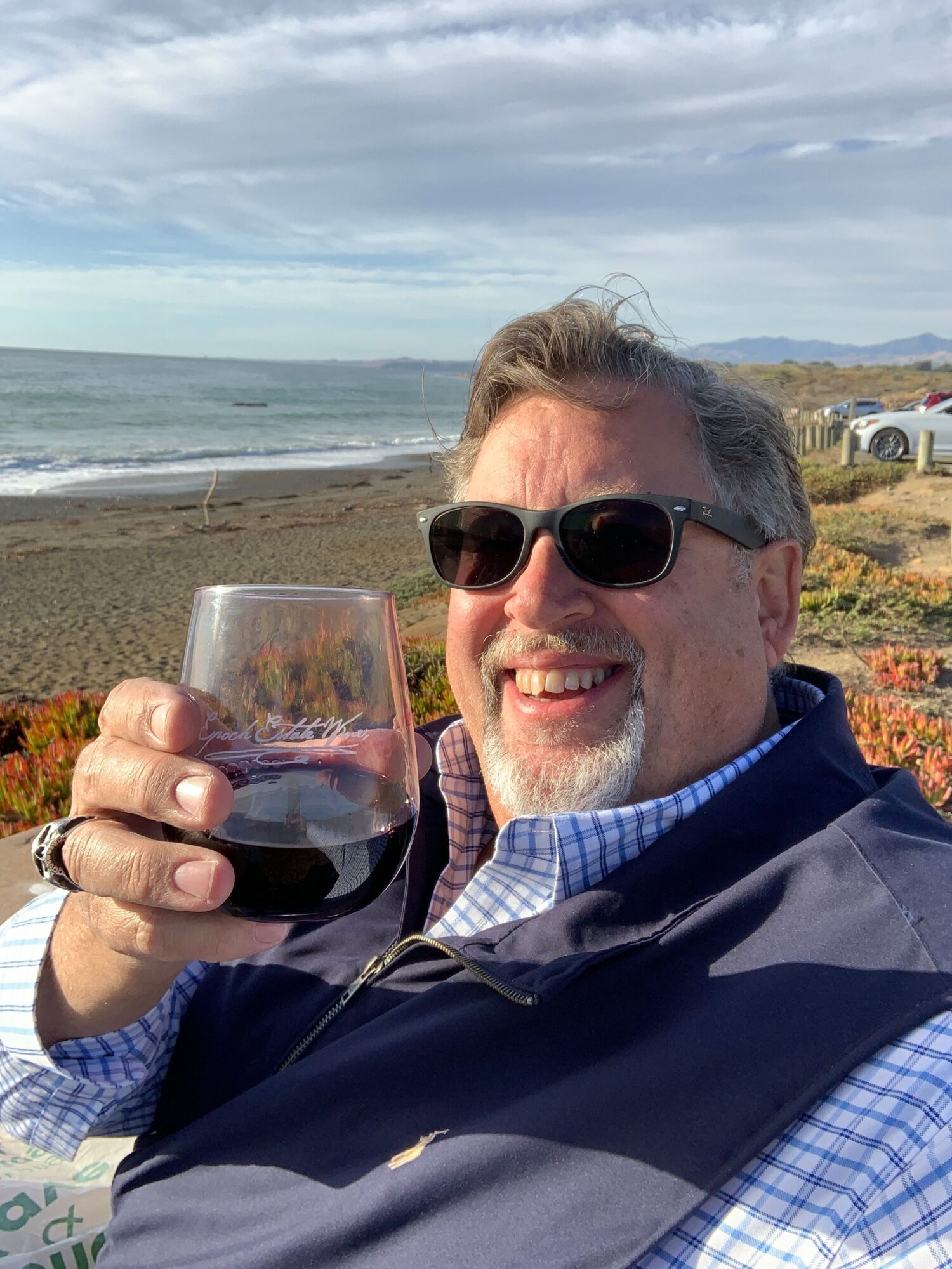 Man with sunglasses smiling at the camera, holding a glass of red wine, on a beach with ocean and sky in background.