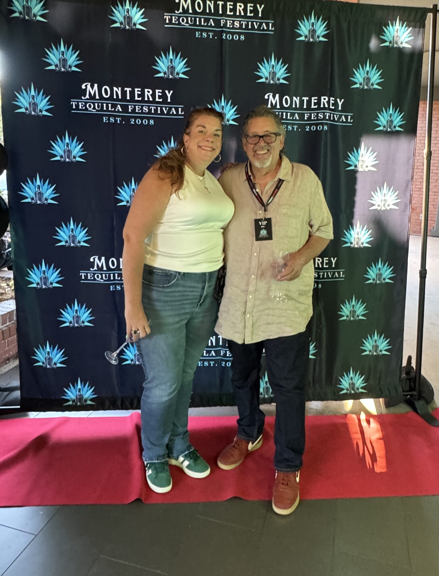Two people standing in front of a Monterey Tequila Festival backdrop, smiling, with a pink carpet beneath them.