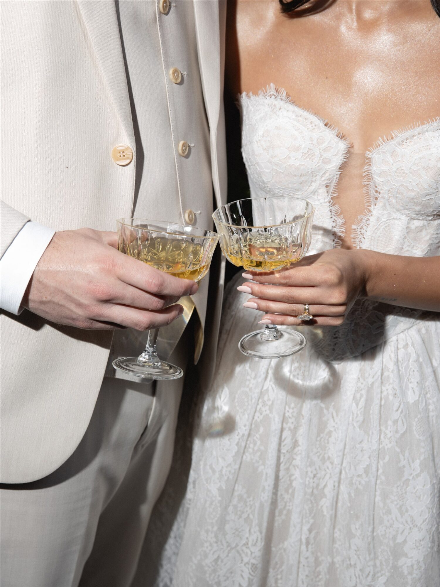Two people in formal attire holding champagne glasses, one in a white suit and the other in a white dress, celebrating.