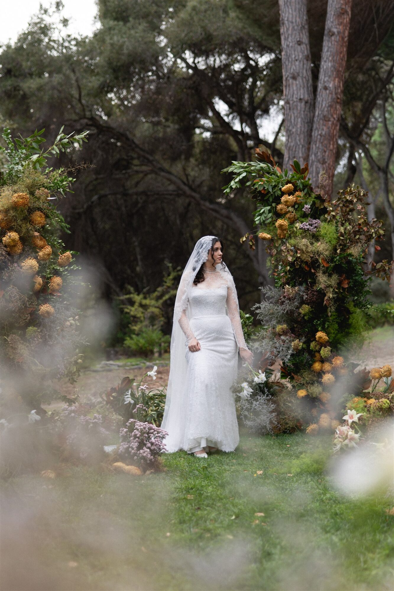 Bride in a white wedding dress and veil standing outdoors among trees and flowers, surrounded by greenery and colorful blooms.