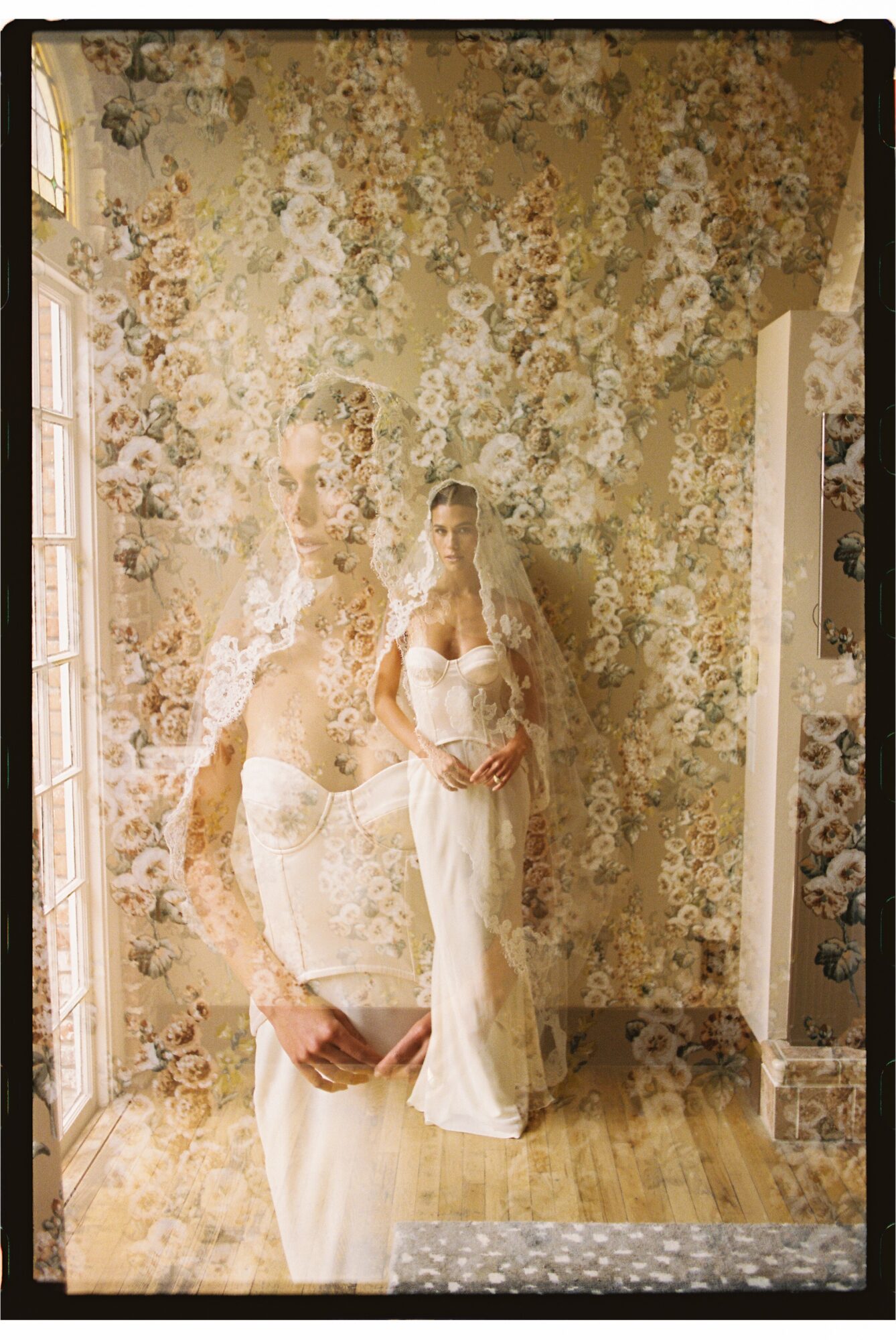 Two women in white dresses standing in a room with floral wallpaper, one holding a bouquet, hardwood floor, mirror reflection.
