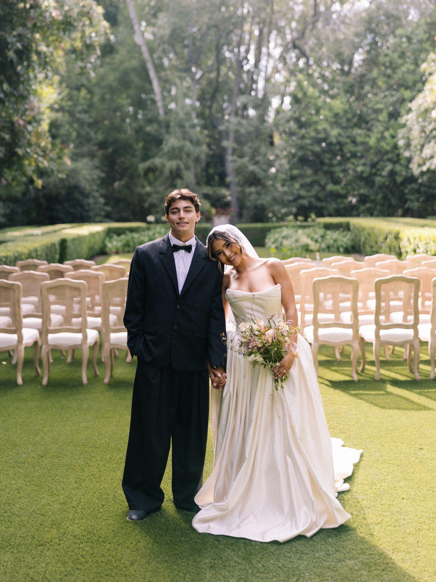 A bride and groom holding hands outdoors on a grassy area with empty chairs behind them, trees in the background.