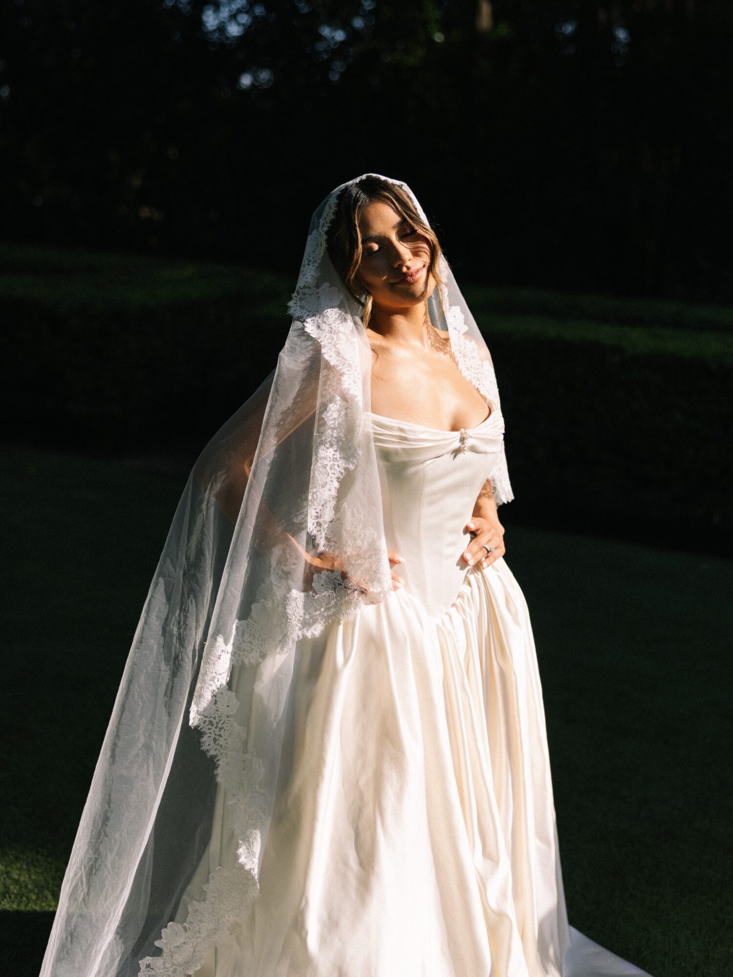 Woman in wedding dress with veil standing outdoors in sunlight, dark background, smiling, facing forward.