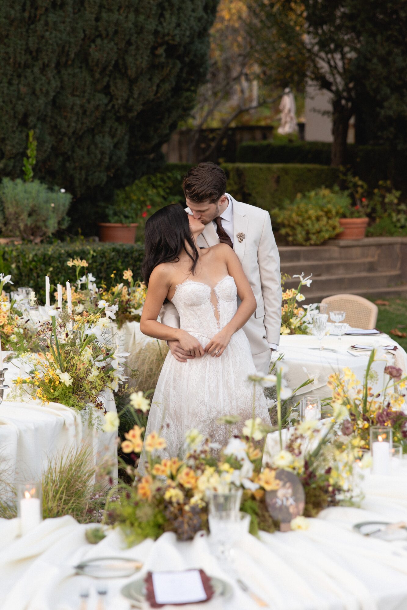 Couple in wedding attire standing outdoors among flowers and tables, embracing each other.