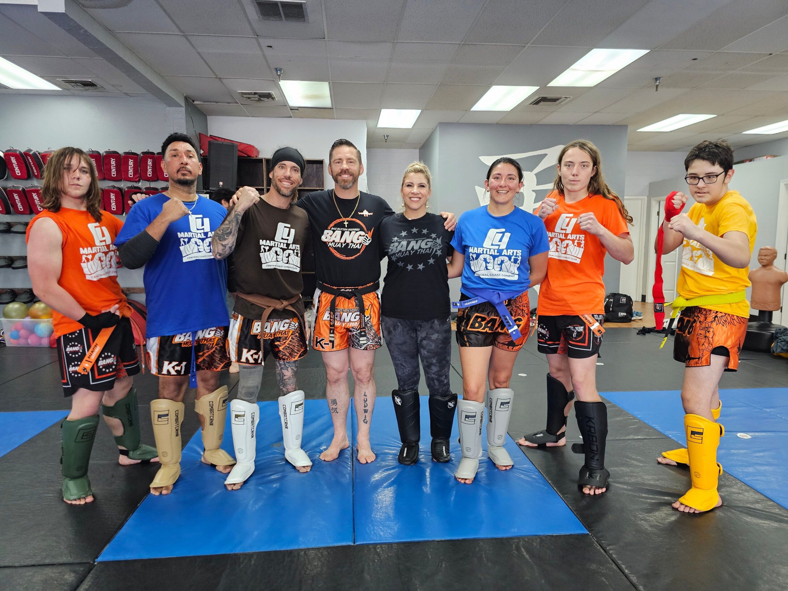 Group of eight people in martial arts uniforms and casual clothing standing in a gym, smiling, with arms crossed or raised.