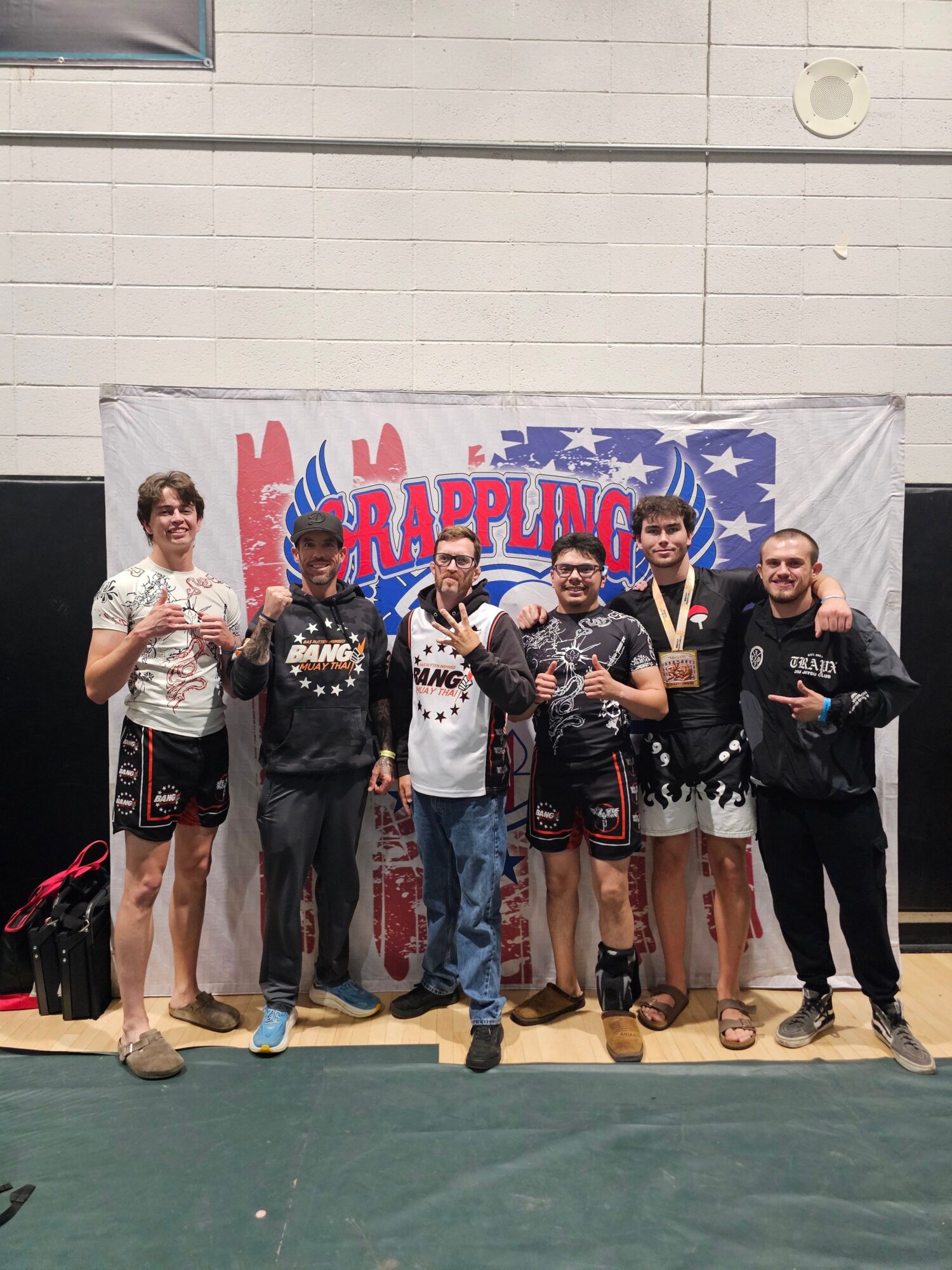 Group of six people standing in front of a banner, some holding medals, indoors, smiling, casual sportswear, gym floor.
