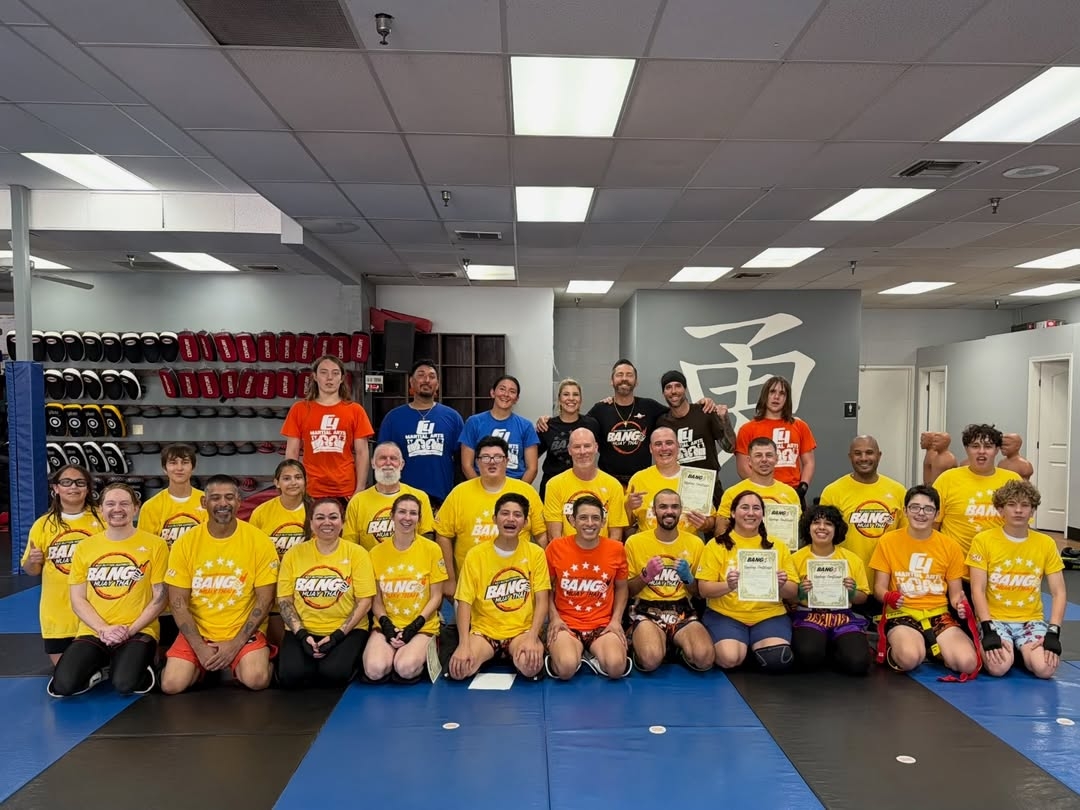 Group of children and adults in a gym, some wearing yellow and orange shirts, posing for a photo.
