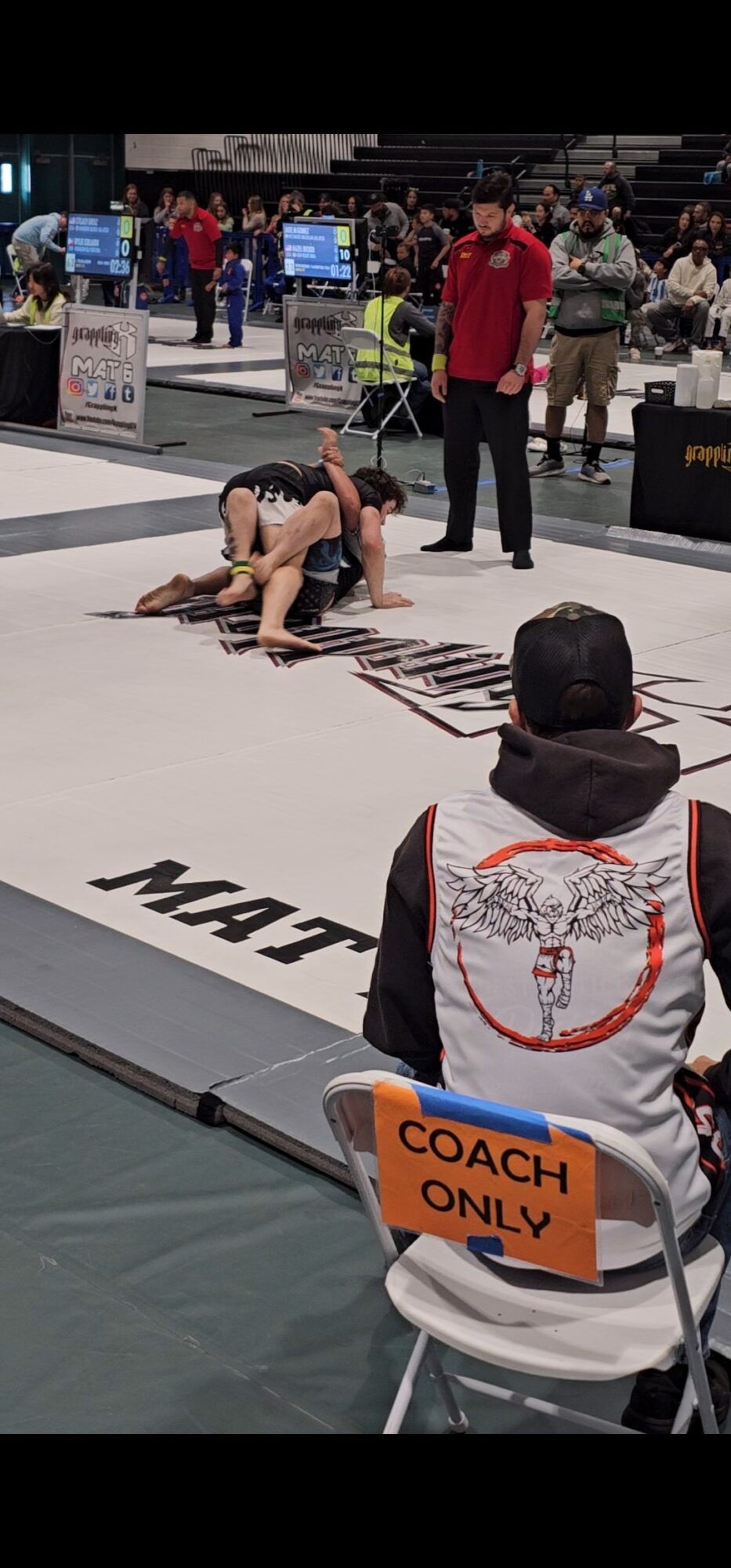 Two athletes grappling on a mat during a competition, with a referee and coach nearby, in an indoor sports arena.