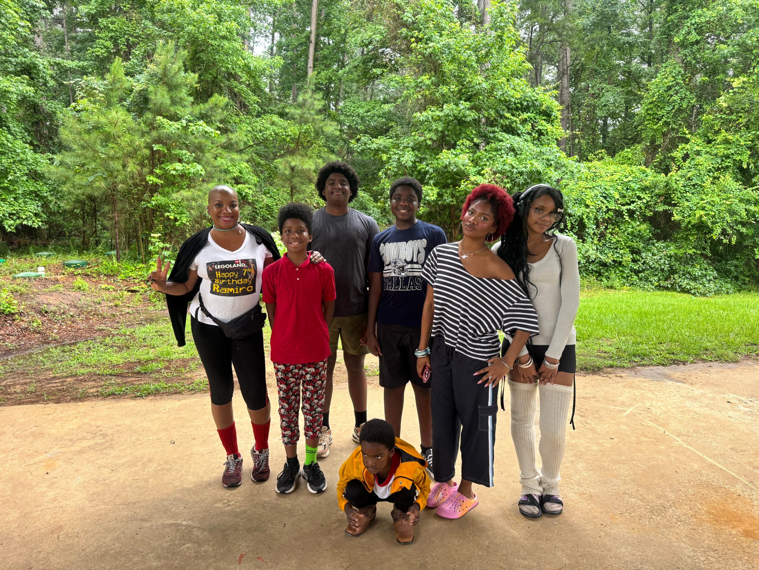 Group of six people standing outdoors on a dirt path with green trees in the background.