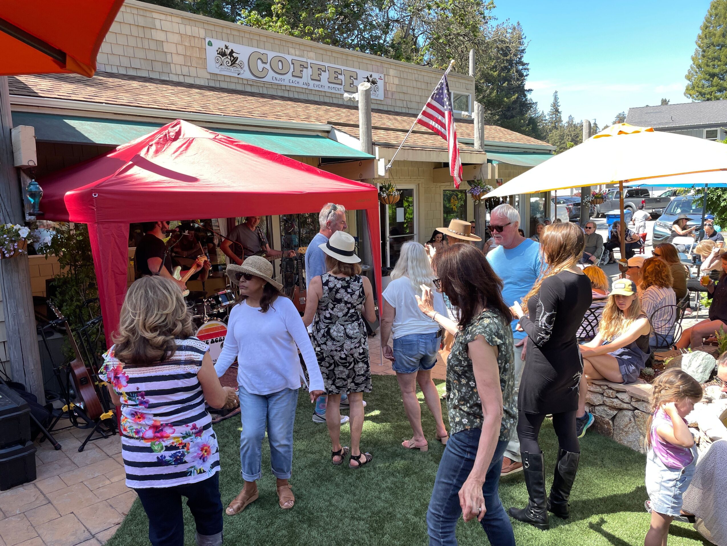 People gather outside a coffee shop with red and yellow tents, some wearing hats, in a sunny outdoor setting.