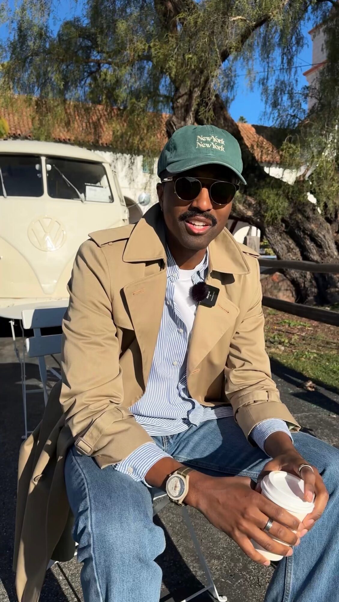Man wearing sunglasses, a cap, beige jacket, and jeans, sitting outdoors holding a drink, with trees and a vehicle in background.