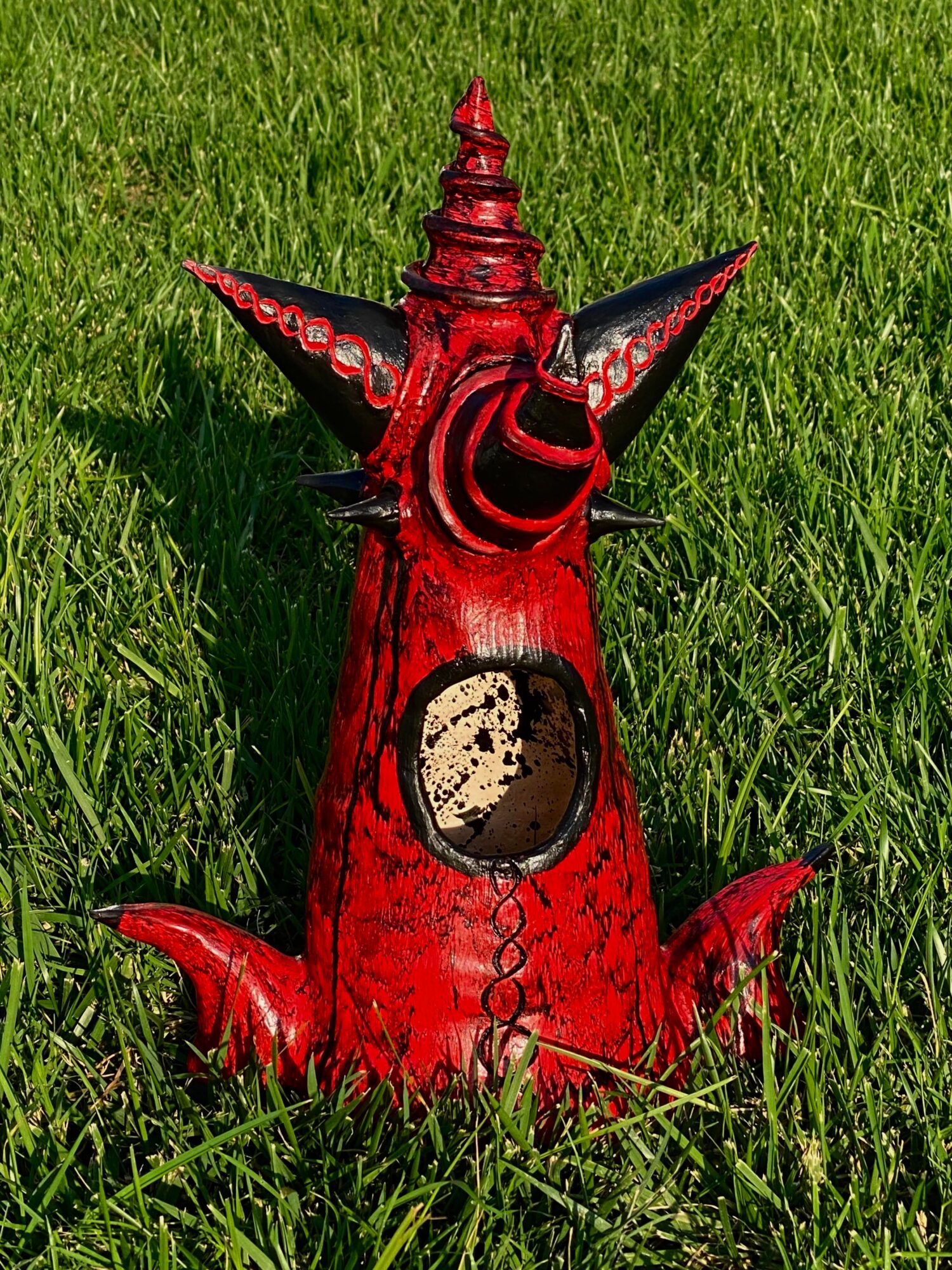 Red, abstract sculpture with pointed elements, set on green grass, featuring a circular opening and textured surface.
