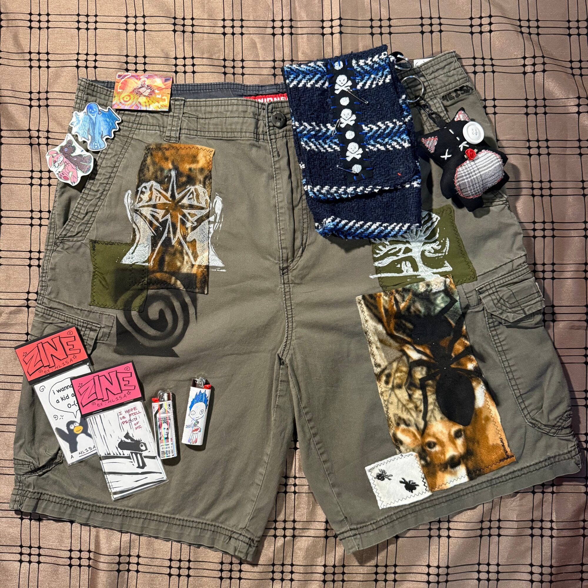 Olive green cargo shorts with various patches, stickers, and small plush toys attached, laid on a checkered fabric background.