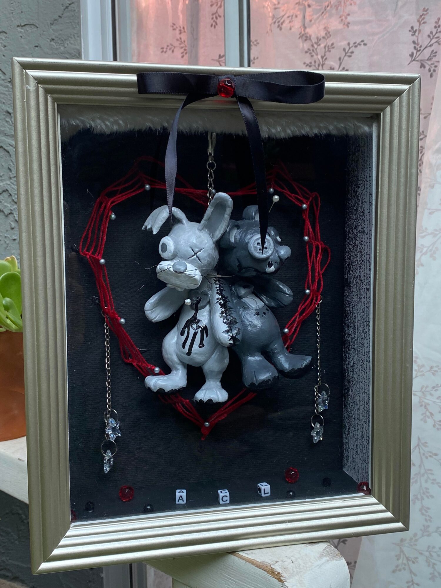 Decorative display with two bunny figurines, red heart-shaped frame, black ribbon, and small objects inside a shadow box.