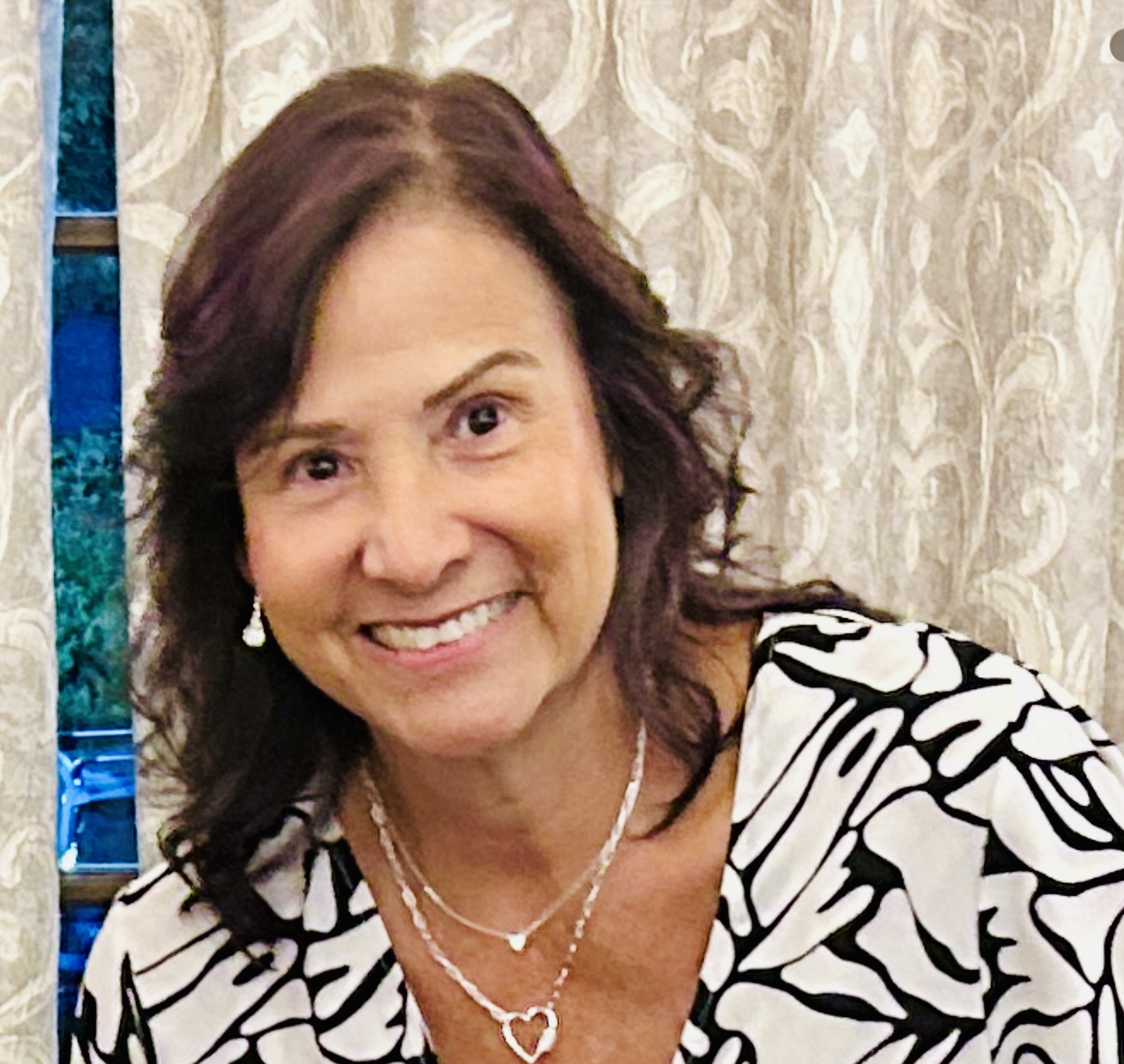 Woman with shoulder-length dark hair smiling, wearing a patterned top and jewelry, in front of curtains.