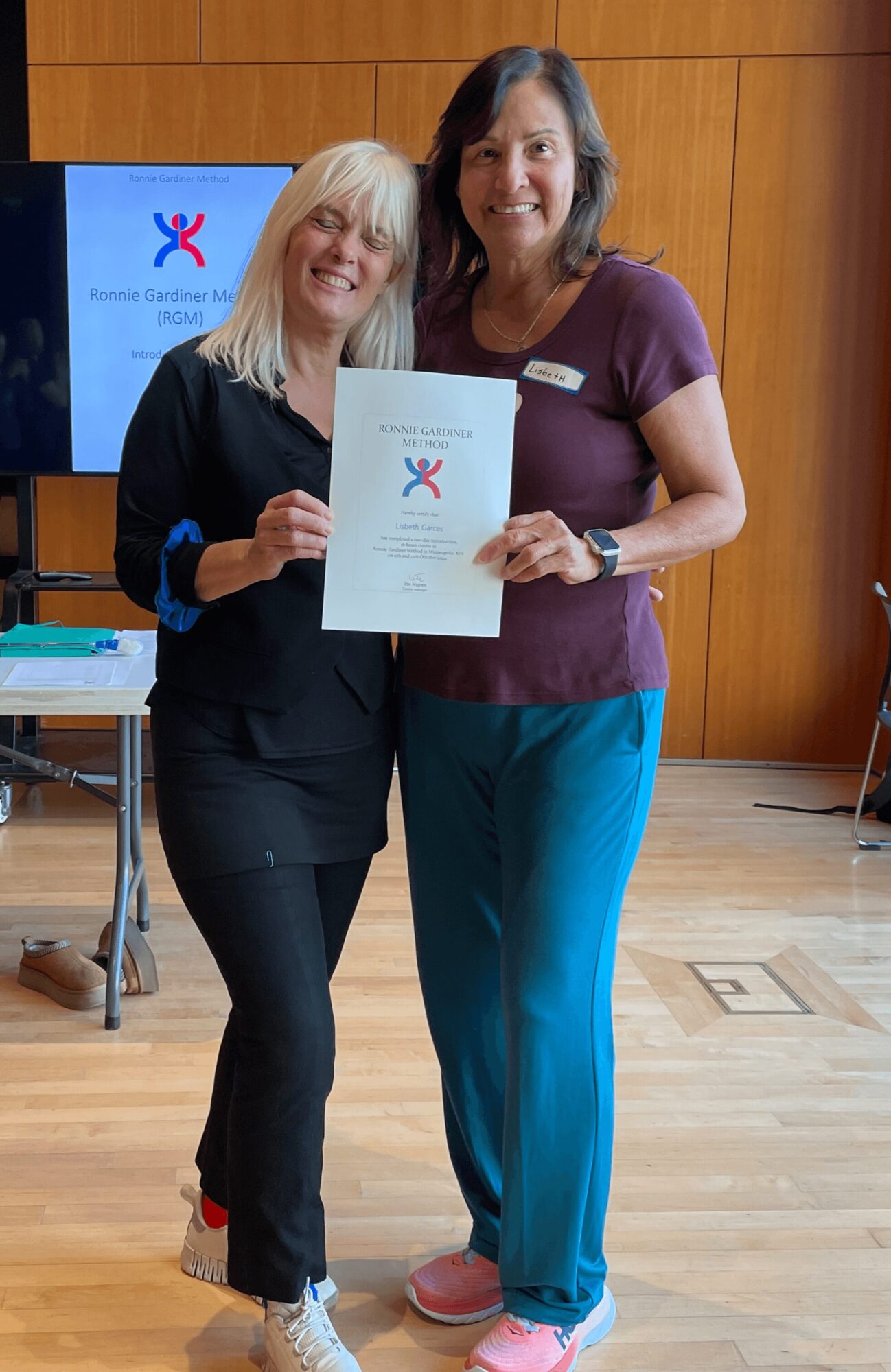 Two women standing indoors, holding a certificate, smiling, with a wooden wall and a screen in the background.