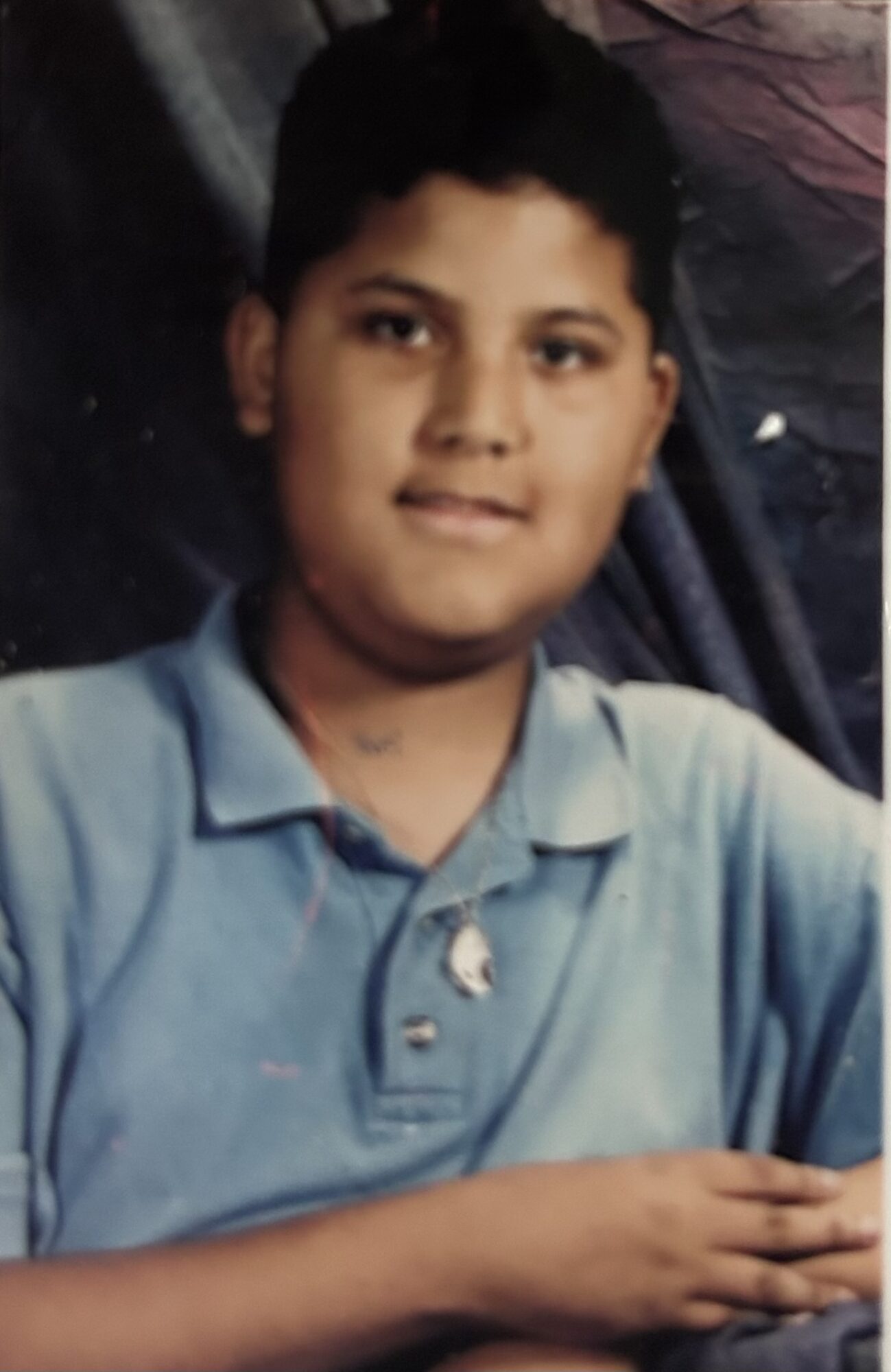 Young boy with short dark hair wearing a light blue polo shirt, sitting with arms crossed, smiling slightly.