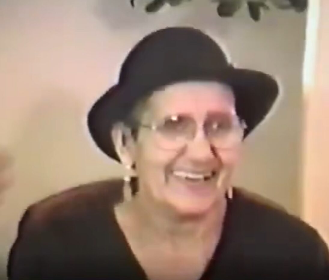 Smiling elderly woman wearing glasses, a black hat, and earrings, indoors with a neutral background.
