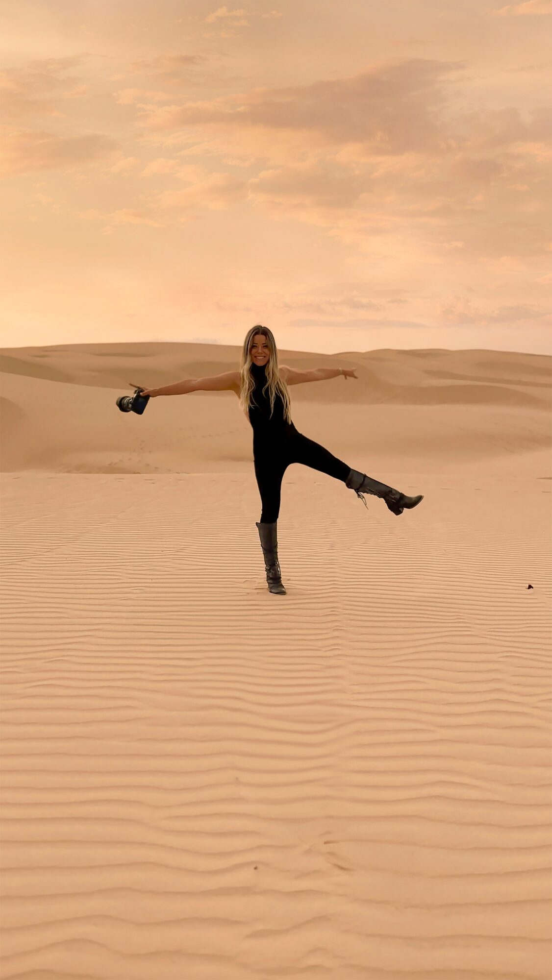 Person standing on sand dunes with arms and leg extended, desert landscape, sunset sky, warm colors